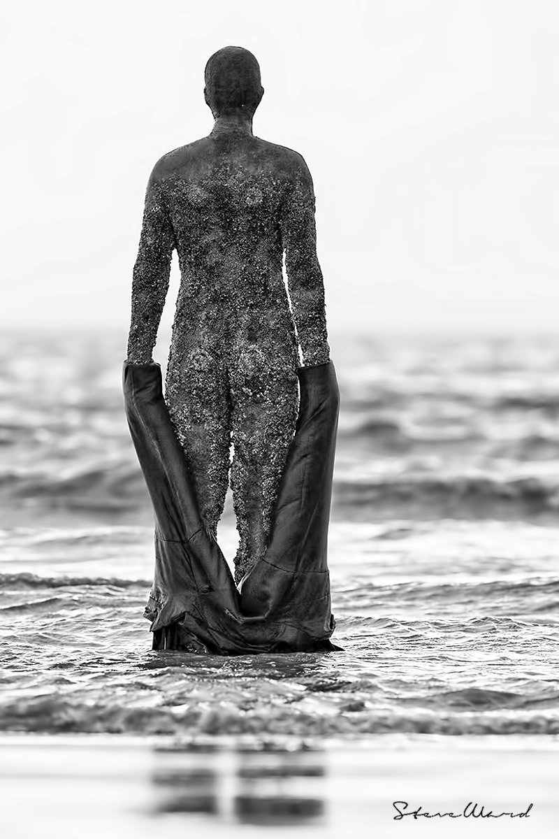 Black and white photo of a person standing in the water, facing away, with their body covered in salt or snow and holding large boots.