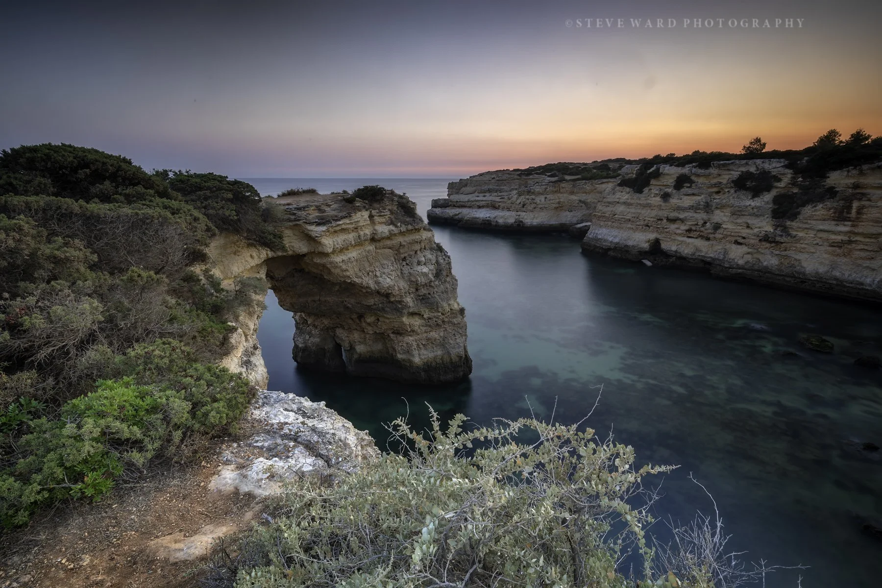 Scenic coastal view of rock formations and a small cove at sunset, with the sky showing pink and purple hues and vegetation in the foreground.