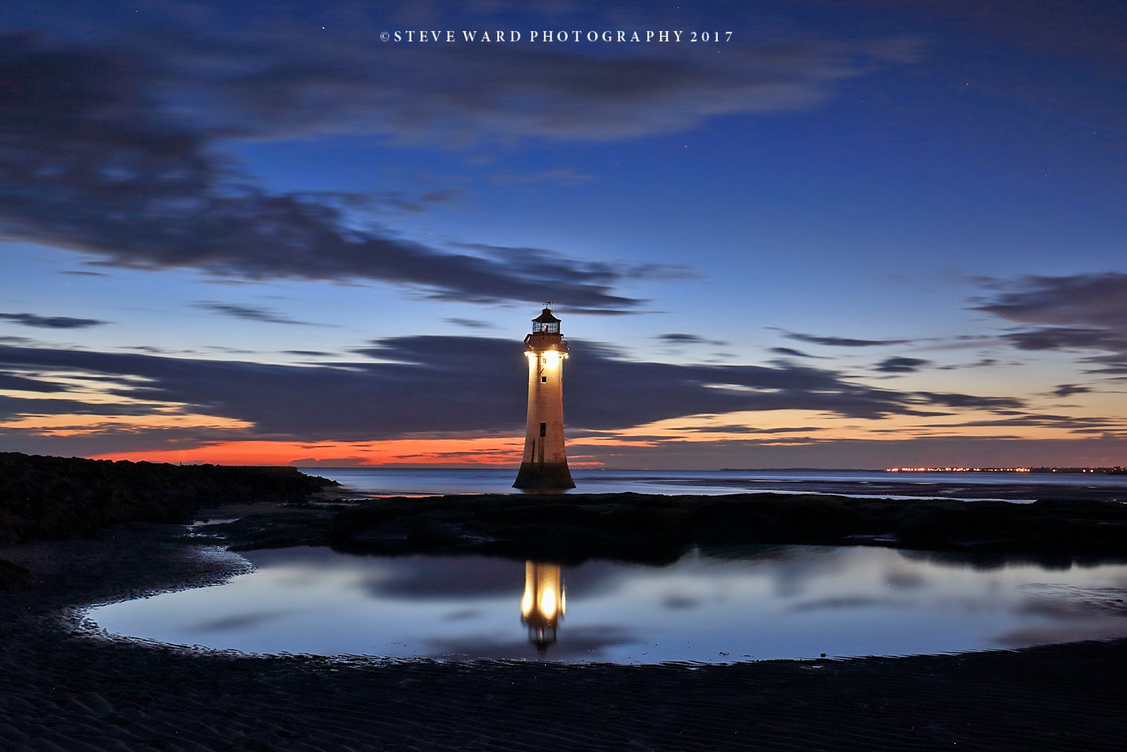 A lighthouse standing in water during twilight, with clouds in the sky and the light from the lighthouse reflecting on the water.
