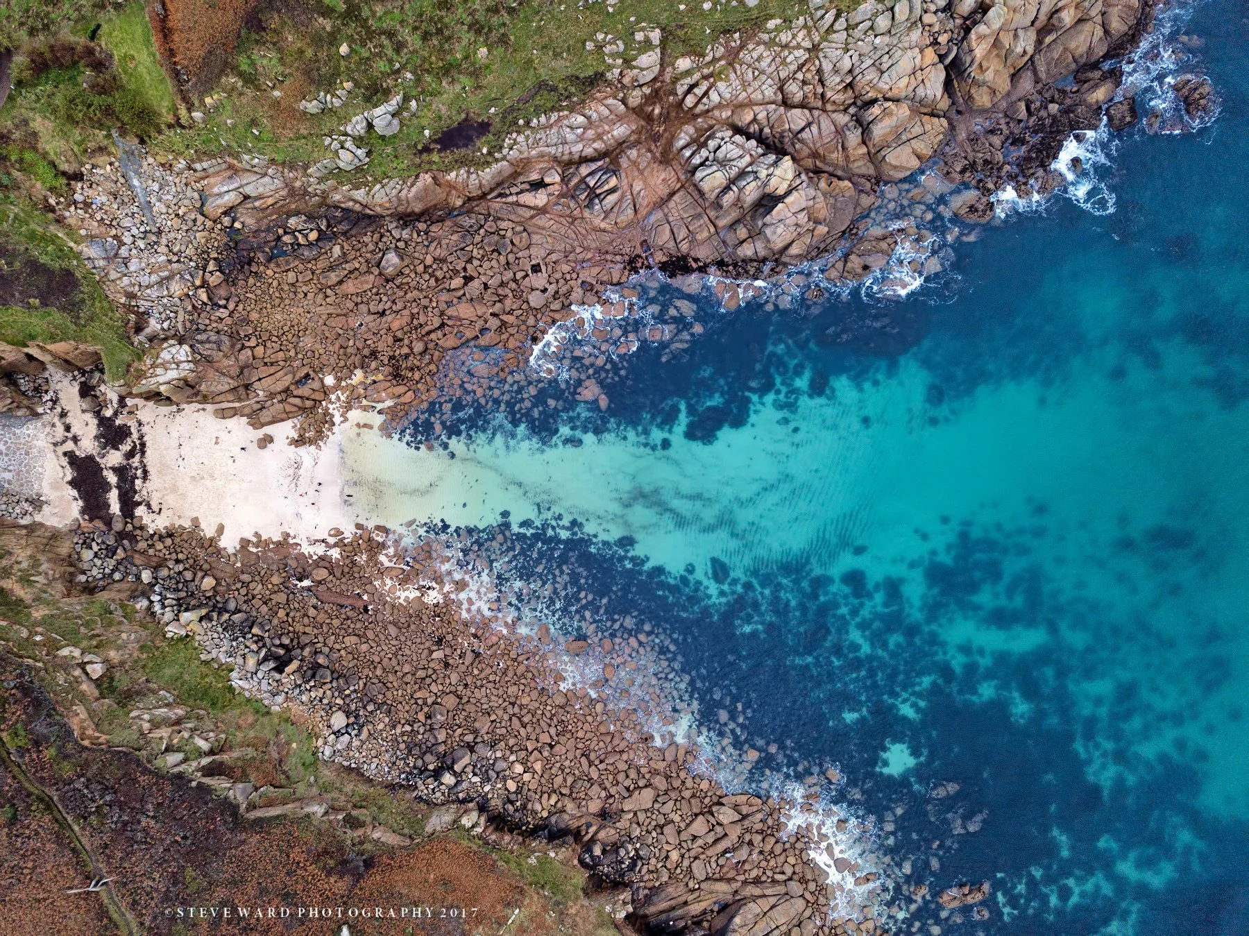 Aerial view of a rocky coastline with clear blue water and a small sandy beach at the bottom left corner.