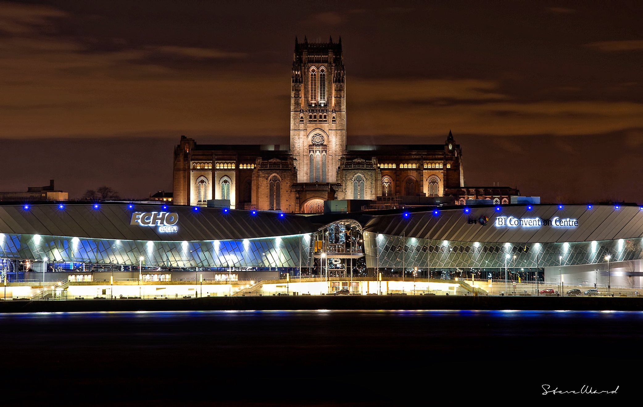 Nighttime photo of the BT Convention Centre and Echo Arena with a historic church in the background, illuminated against a cloudy sky.