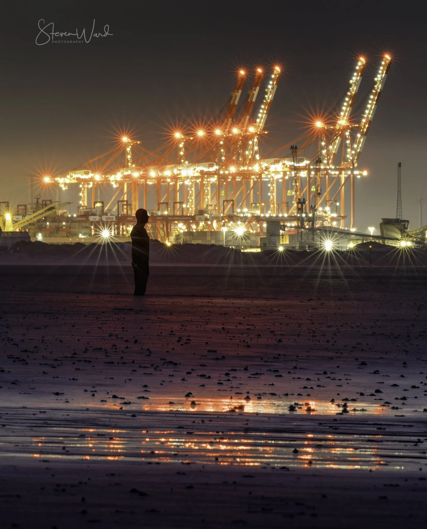 A person standing on a beach at night, looking at brightly lit port cranes and containers in the background.