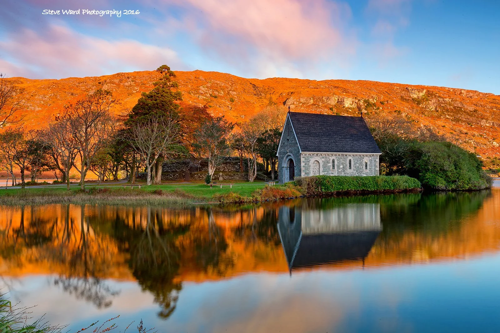 A peaceful scene of a small stone church with a dark roof, surrounded by trees and greenery, reflected in a calm body of water. In the background, a hill or mountain with a reddish-orange hue under a partly cloudy sky.