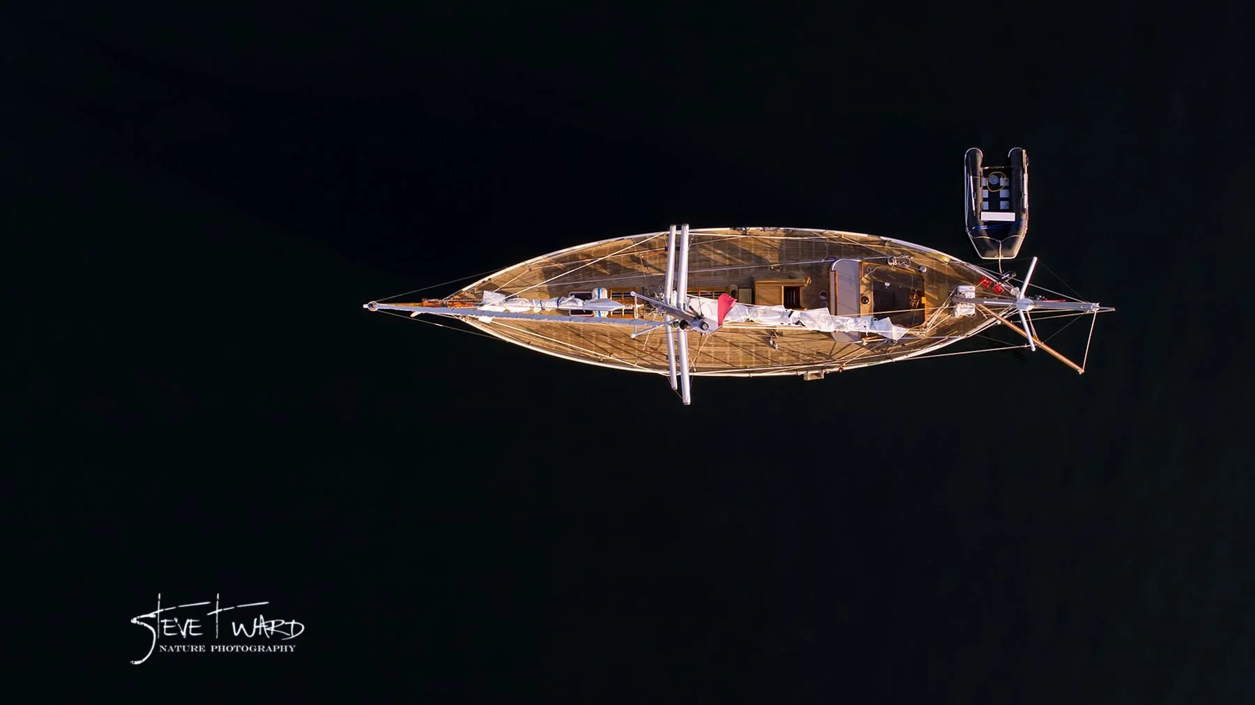 Top-down view of a wooden sailboat on dark water, with a small motorboat nearby, taken during daylight.