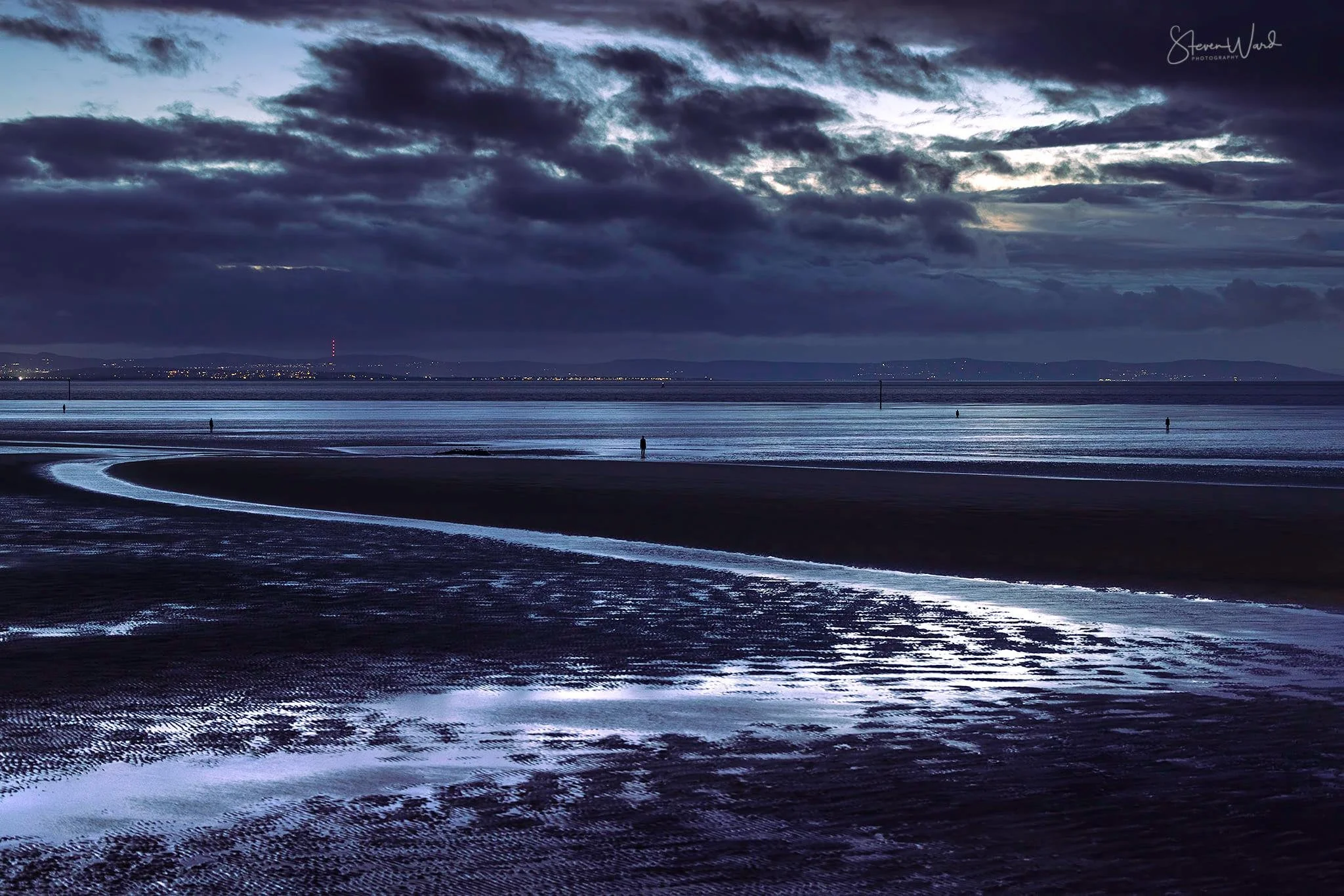 A dark, cloudy sky over a beach with a winding creek reflecting moonlight, and a person standing alone in the distance.
