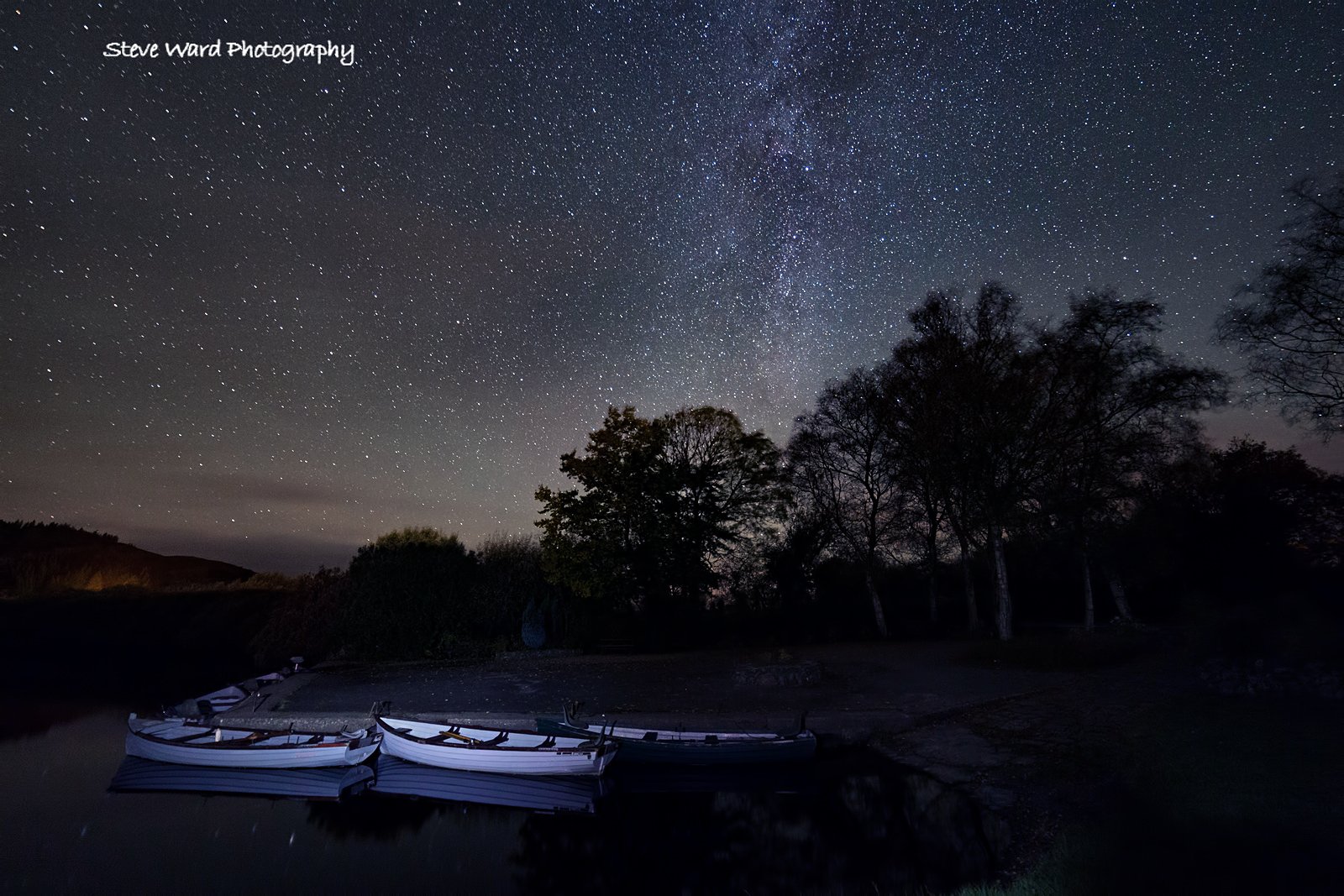 Night sky filled with stars and the Milky Way, with a silhouette of trees and several boats on calm water in the foreground.