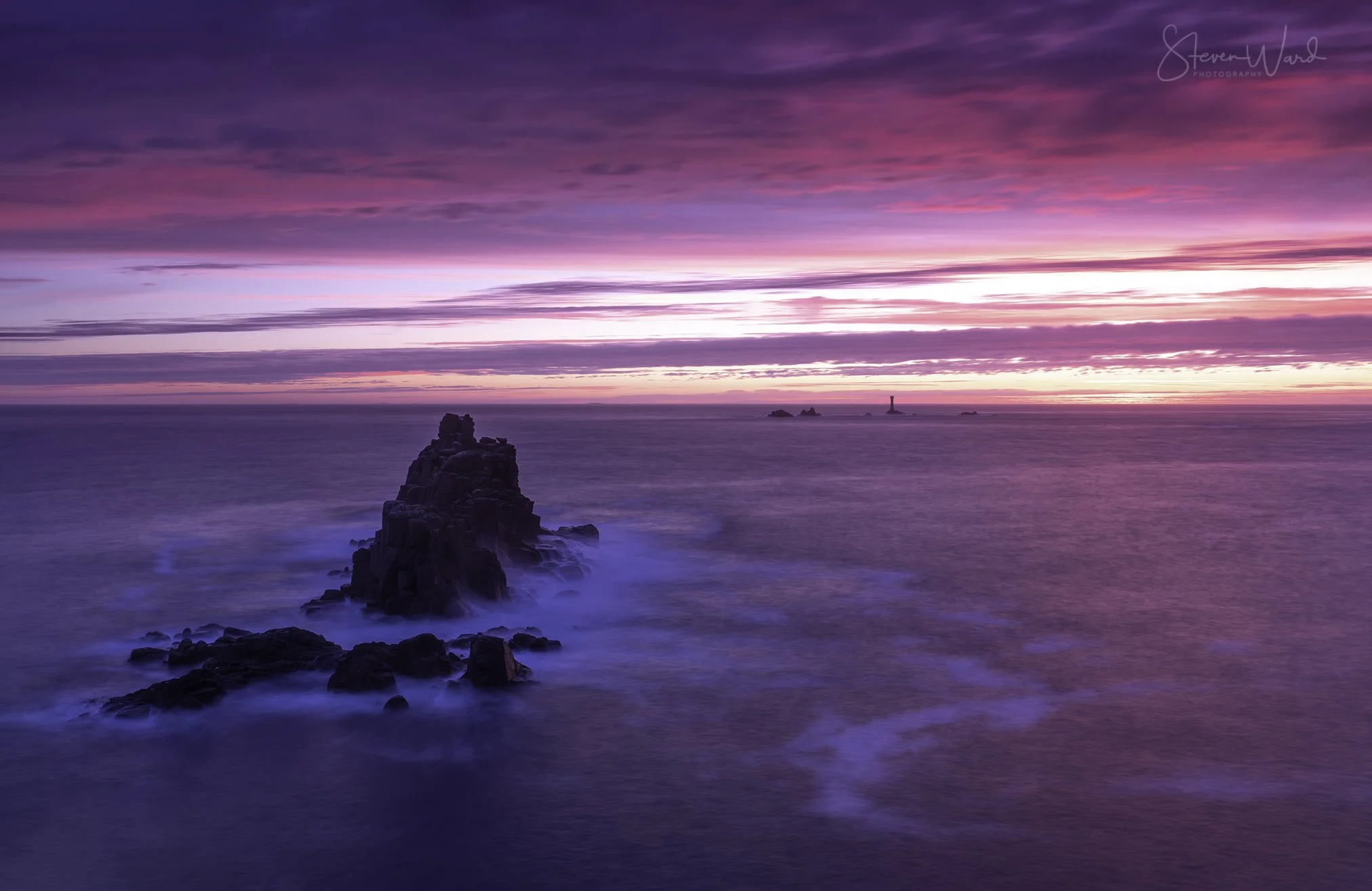 A colorful sunset over the ocean with purple, pink, and orange hues in the sky. Rocky formations extend into the water, with a distant lighthouse on a small island.
