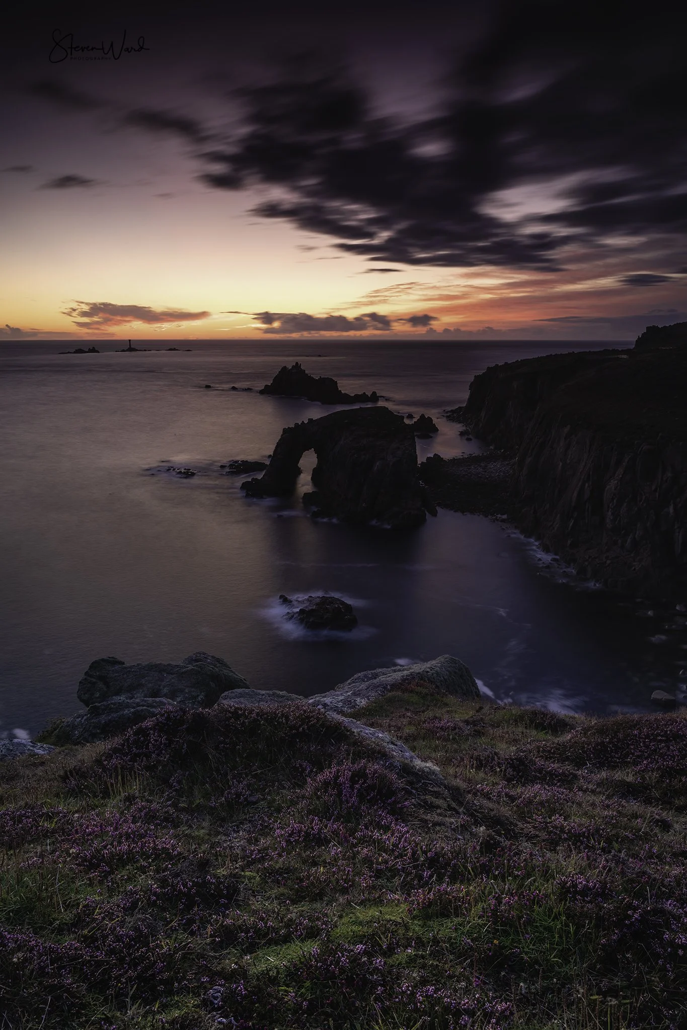 Scenic view of a coastal landscape during sunset with purple and orange hues in the sky, rocky formations in the water, and purple flowering plants in the foreground.