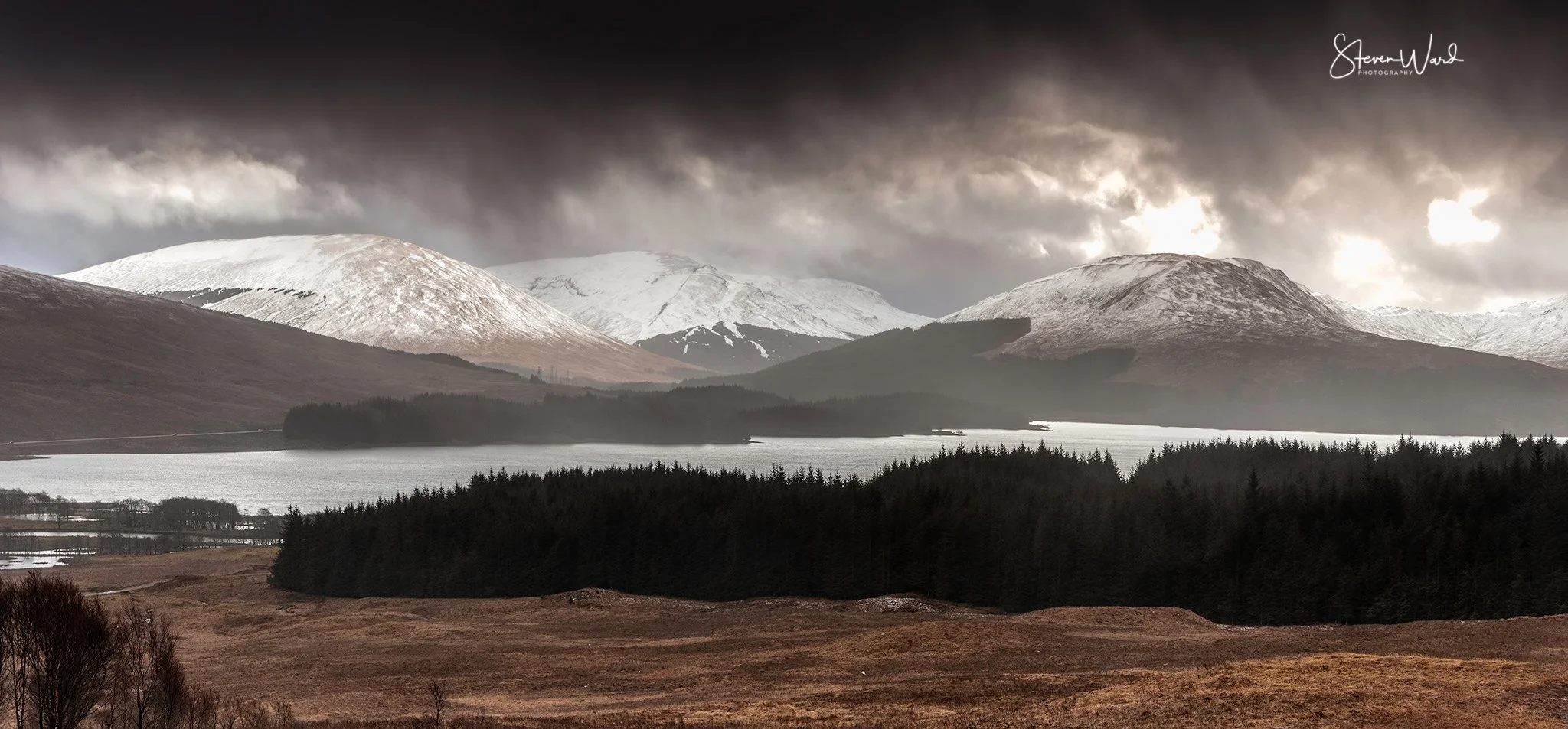 Snow-capped mountains over a lake with dark clouds and a forested foreground in a moody landscape