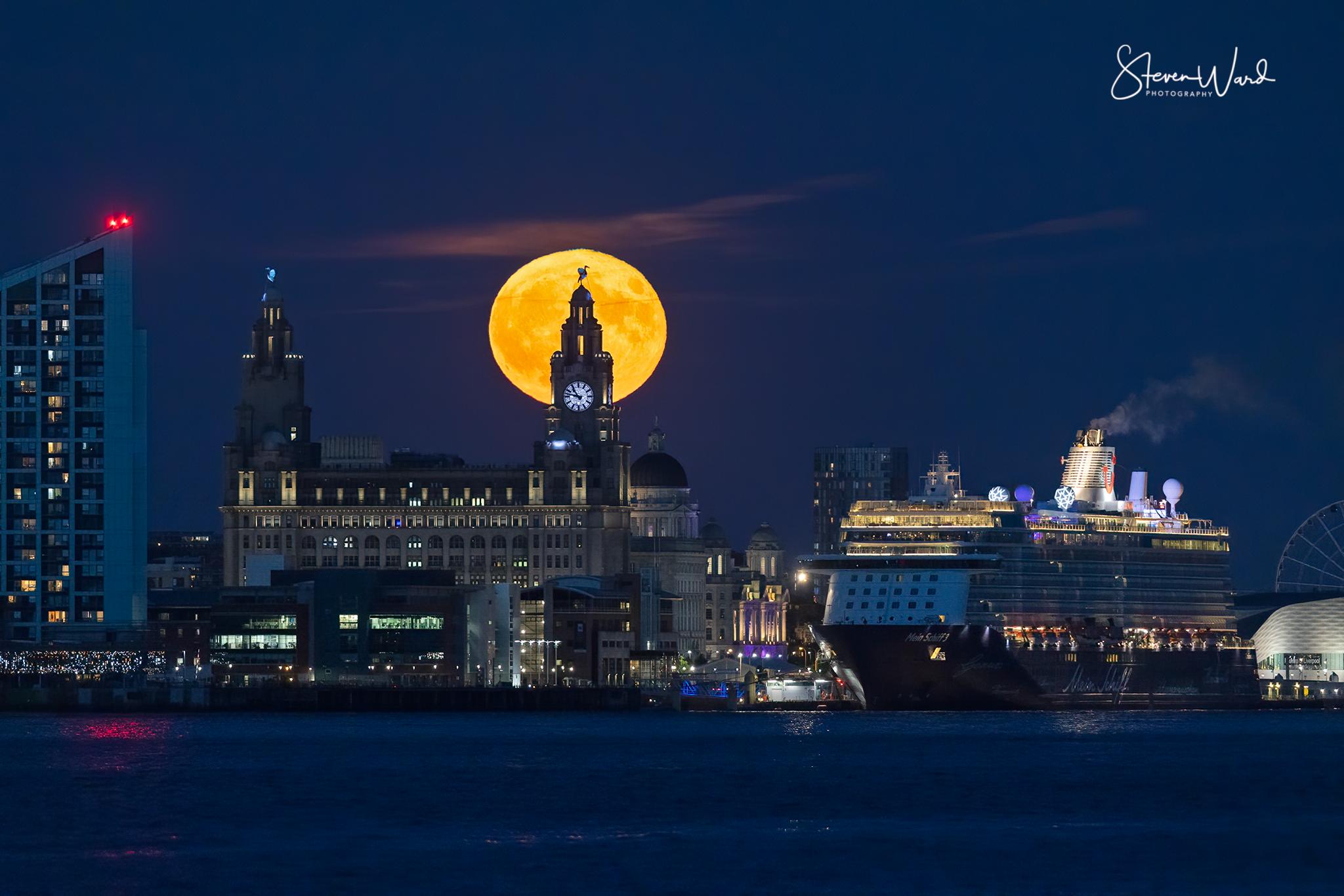 Nighttime city skyline with a large, bright full moon rising behind a historic building with a clock tower, a modern cruise ship docked at the harbor, and various other buildings illuminated.