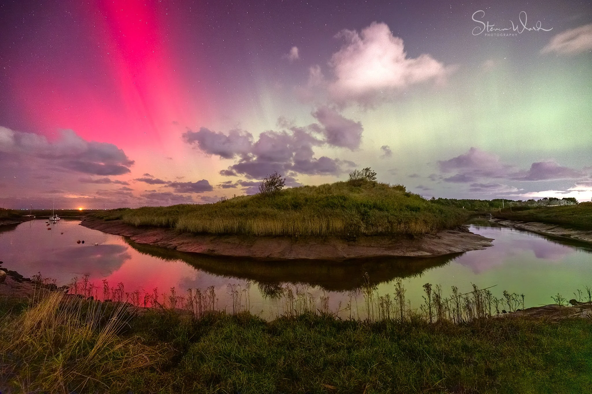 Nighttime landscape featuring a river with a small boat on the left, a grassy hill in the center, and an Northern Lights display with pink and green hues in the sky, along with clouds and a starry night sky.