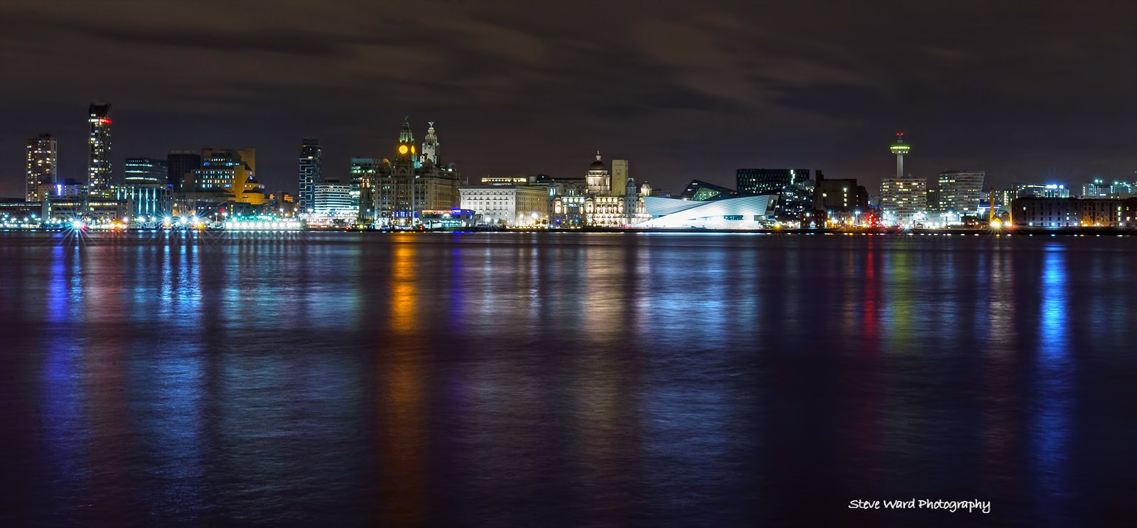 Night skyline of a city reflecting in water with illuminated buildings and a cloudy sky.