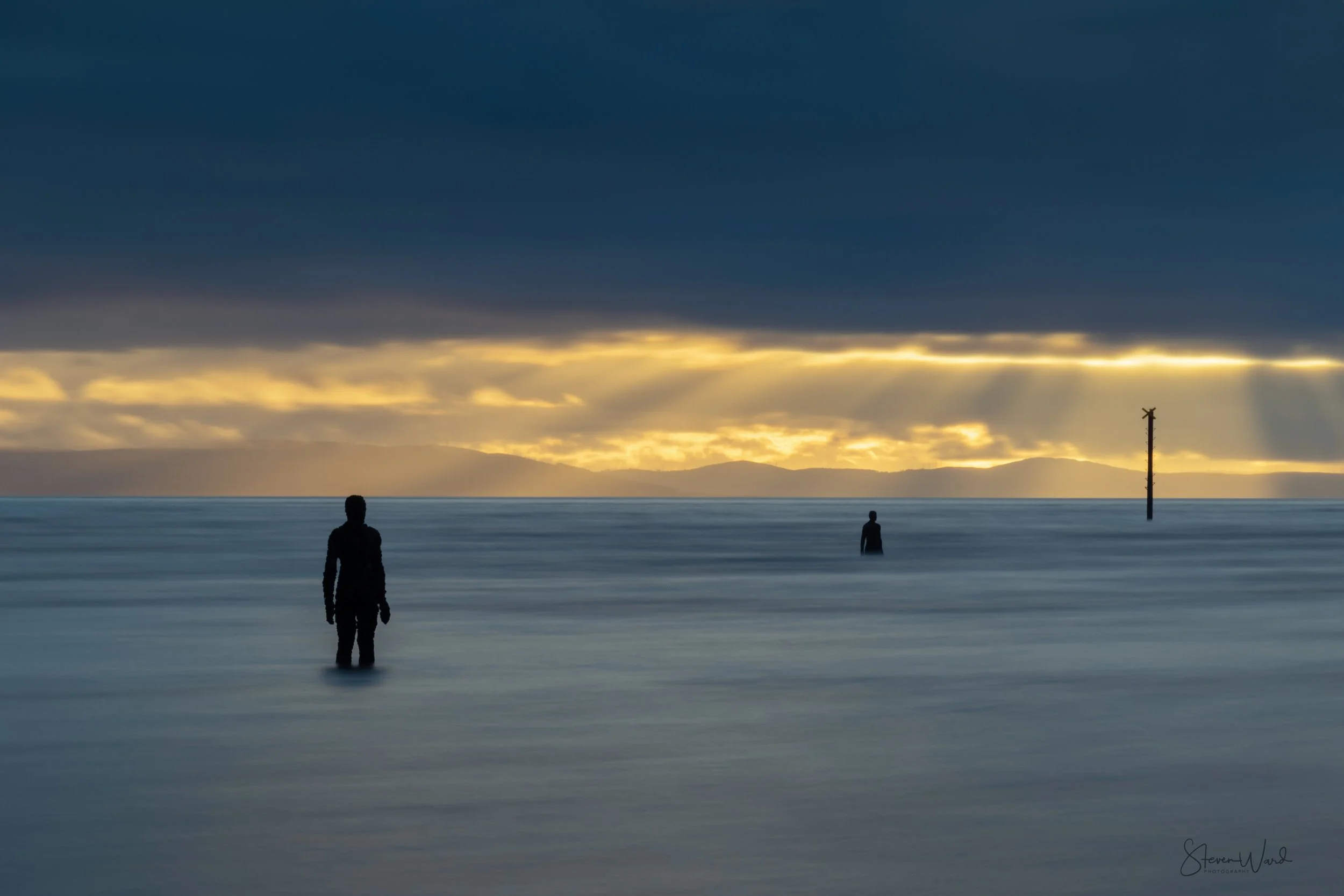 Silhouettes of two people standing in calm ocean waters at sunset with dark clouds and golden sunlight in the sky, and a distant landmass and utility pole visible on the horizon.