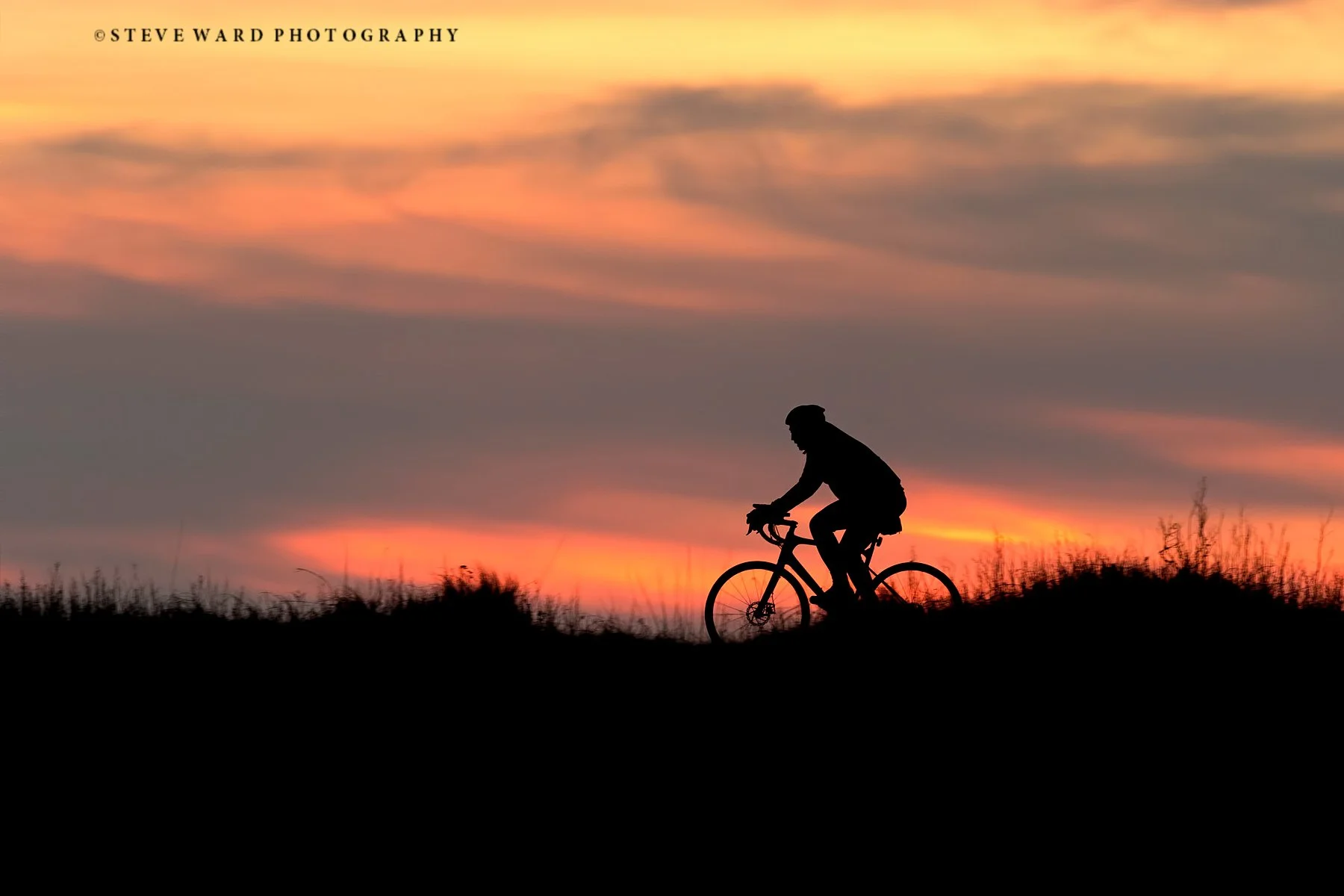 Silhouette of a person riding a bicycle at sunset with colorful sky and clouds in the background.