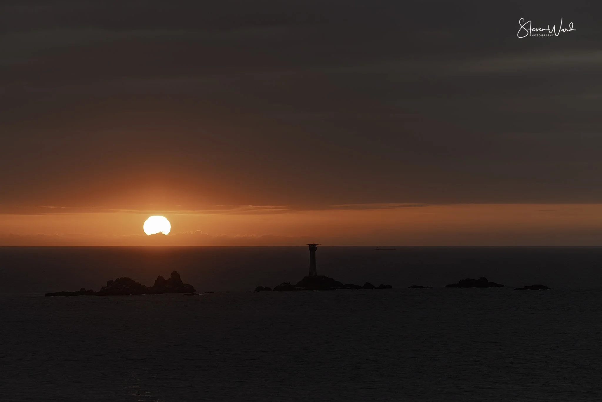 Sunset over the ocean with a lighthouse on a small rocky island and a few smaller rocks visible in the water.