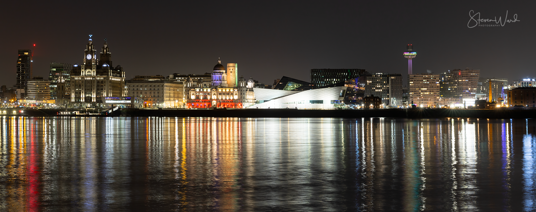 Nighttime city skyline reflecting in the water, with illuminated historic and modern buildings and a tower in the background.