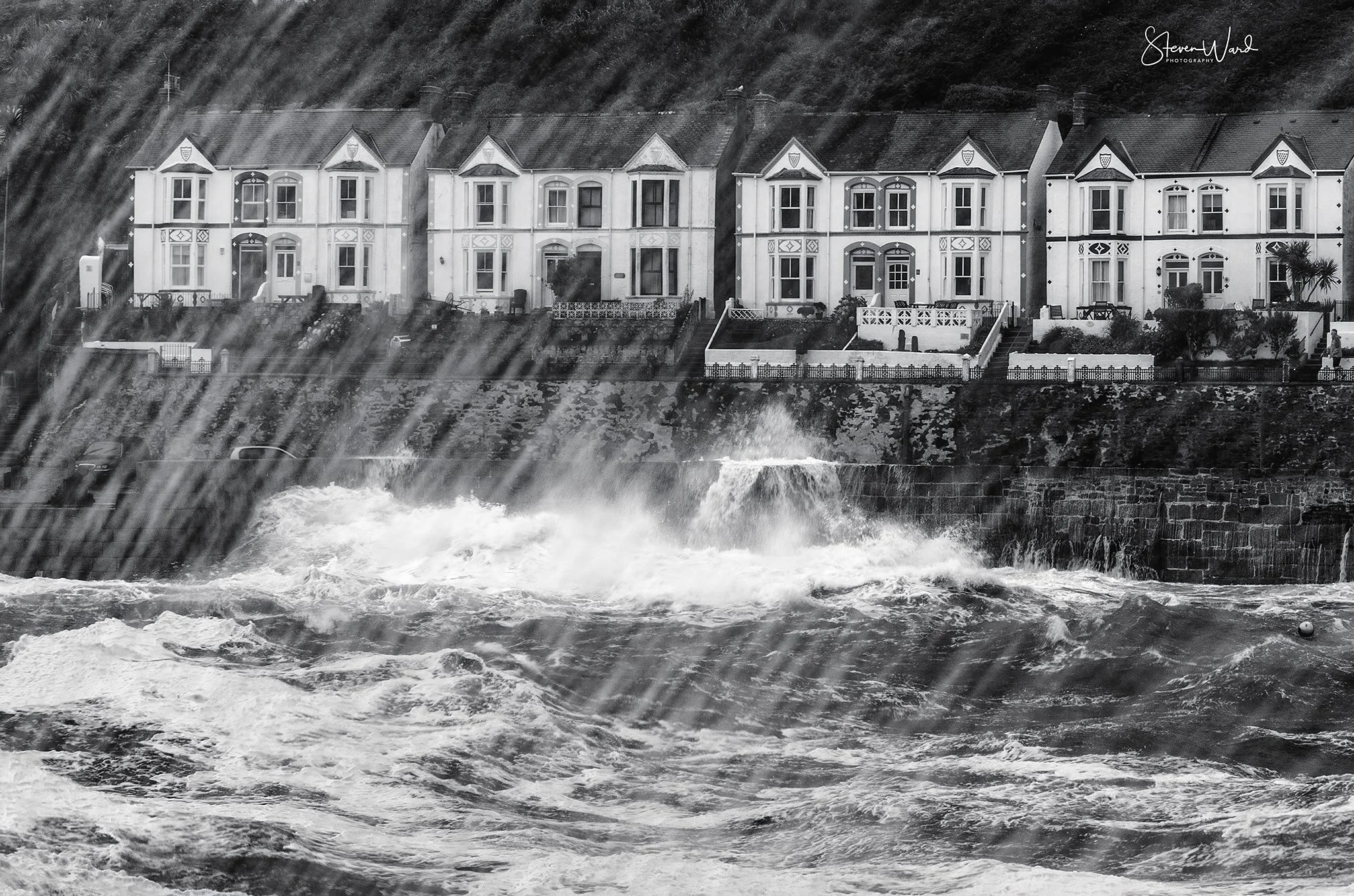A row of white houses on a hillside overlooking ocean waves crashing against a sea wall, with waterfalls splashing over the wall.