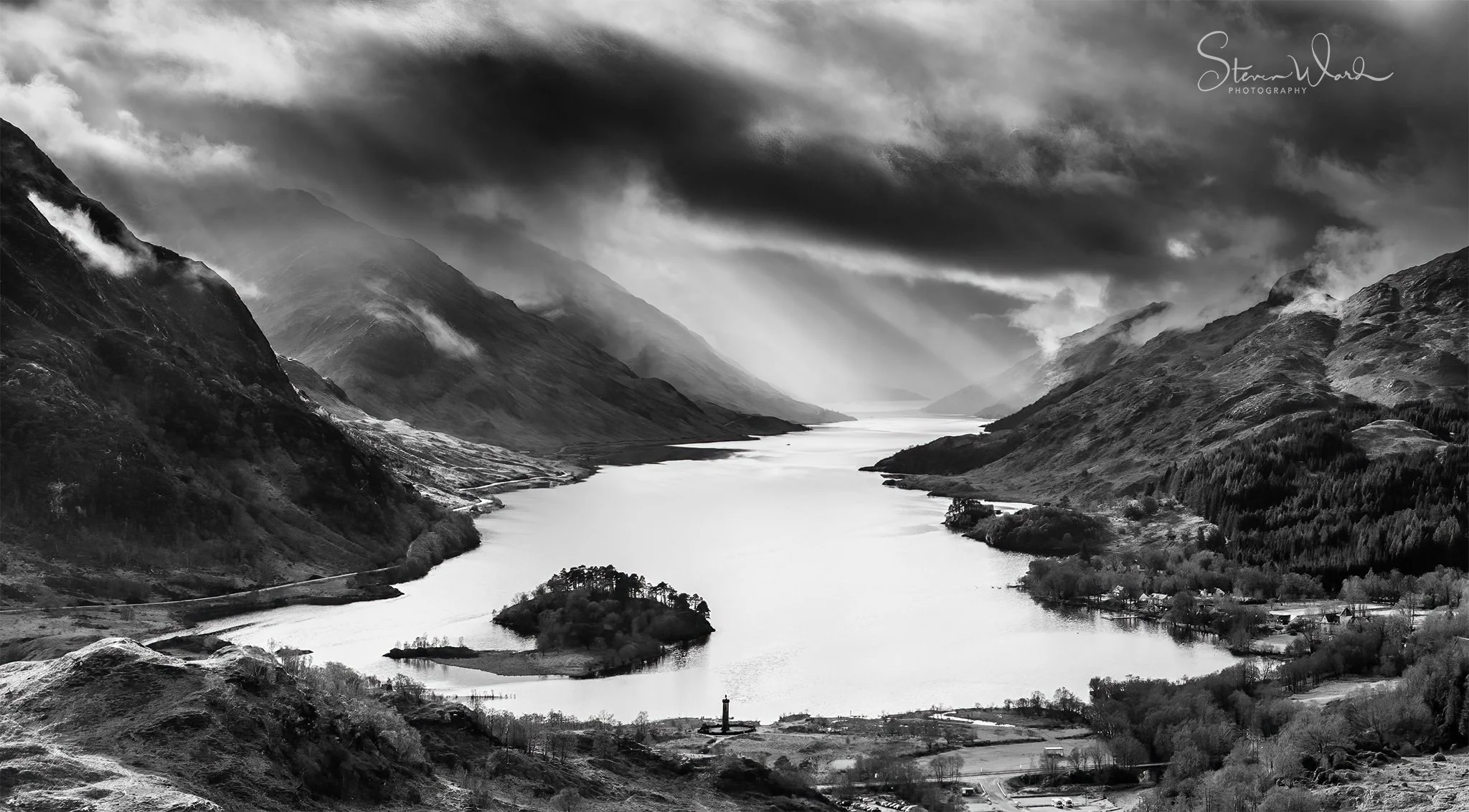 Black and white landscape photo of a river flowing through a valley surrounded by mountains, with cloudy skies overhead.