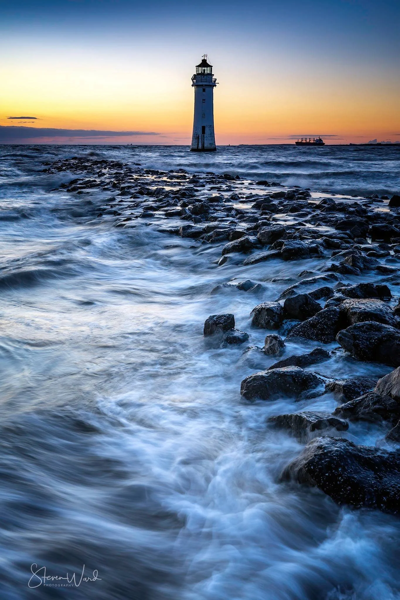 A lighthouse standing in the ocean with waves crashing against rocks, during a sunset or sunrise.