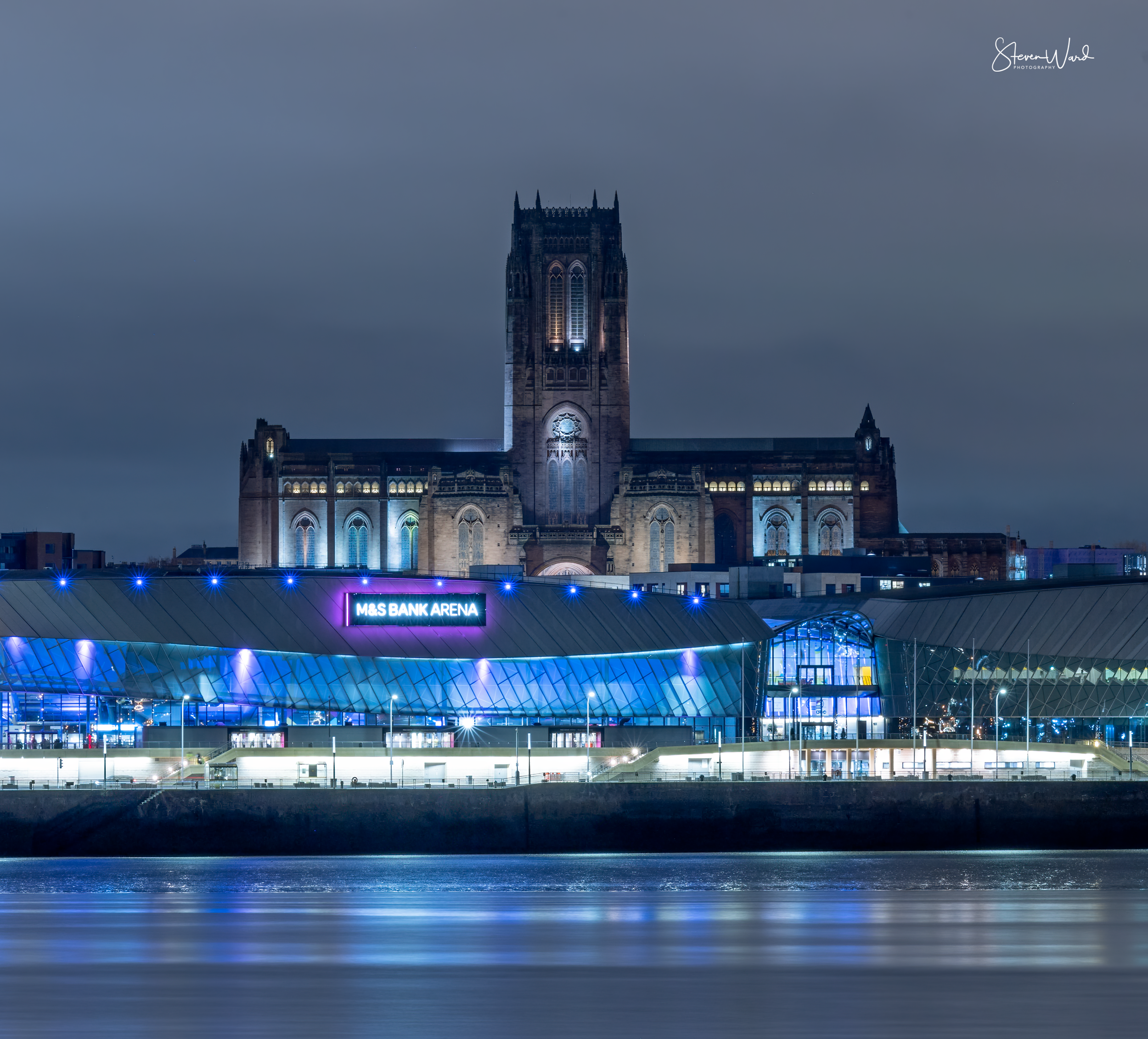 Night view of the M&S Bank Arena with blue lights, with a historic cathedral in the background illuminated in the night sky.