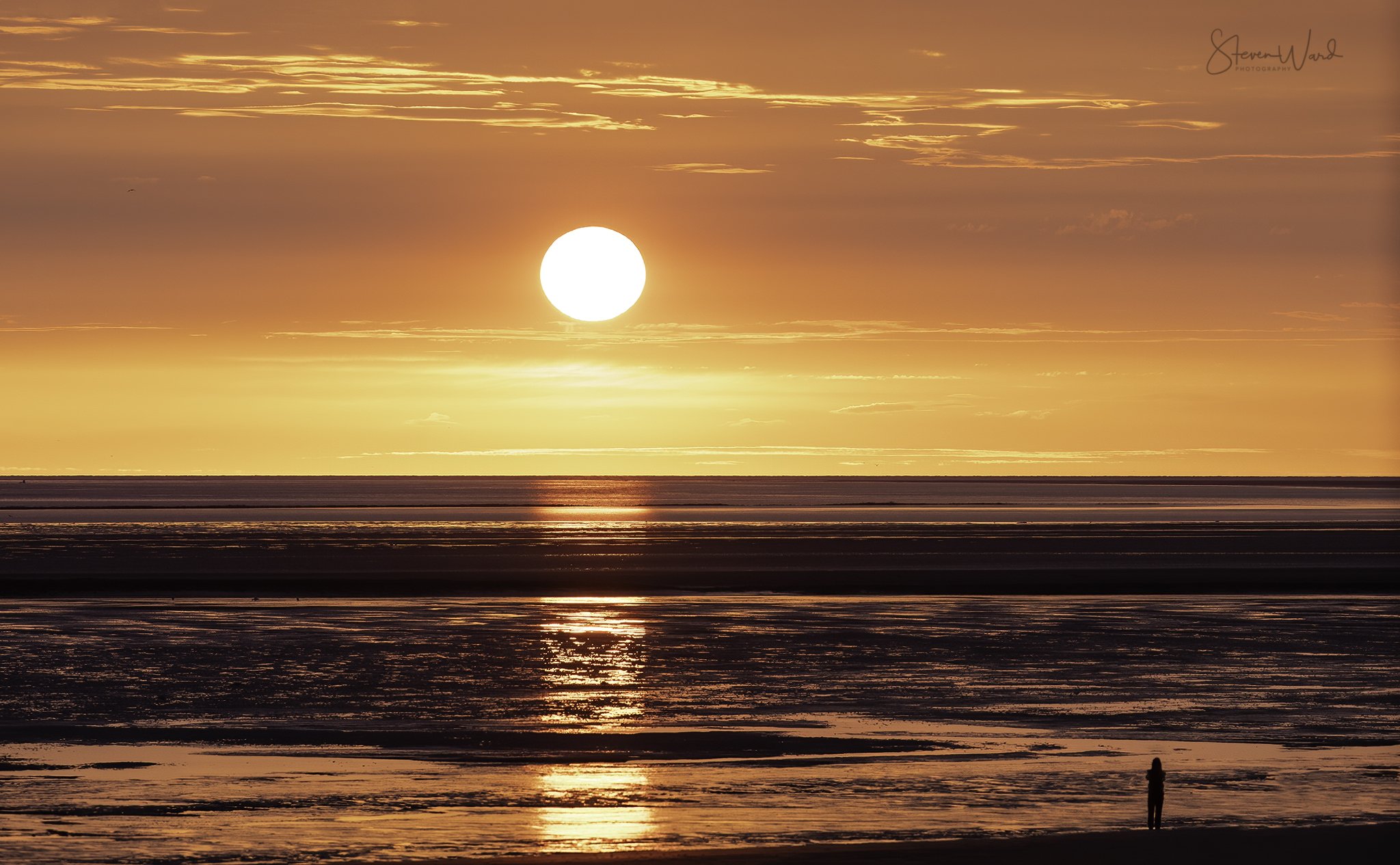 A person standing on a beach during sunset, with the sun low in the sky, casting orange and pink hues across the water and sky.