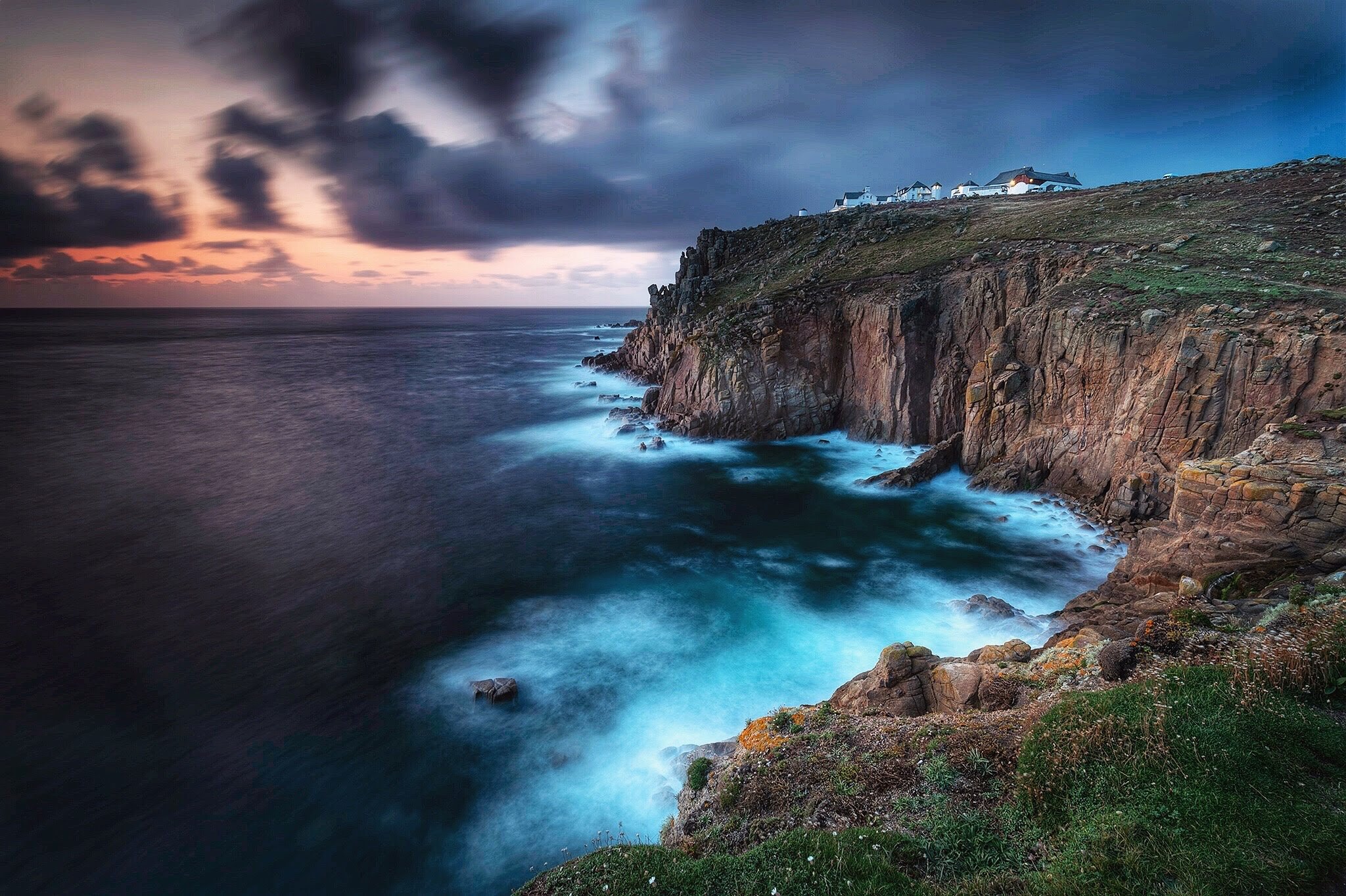 Cliffside coastline at dusk with white buildings on top, rocky cliffs, and ocean waves crashing against the rocks under a cloudy sky.