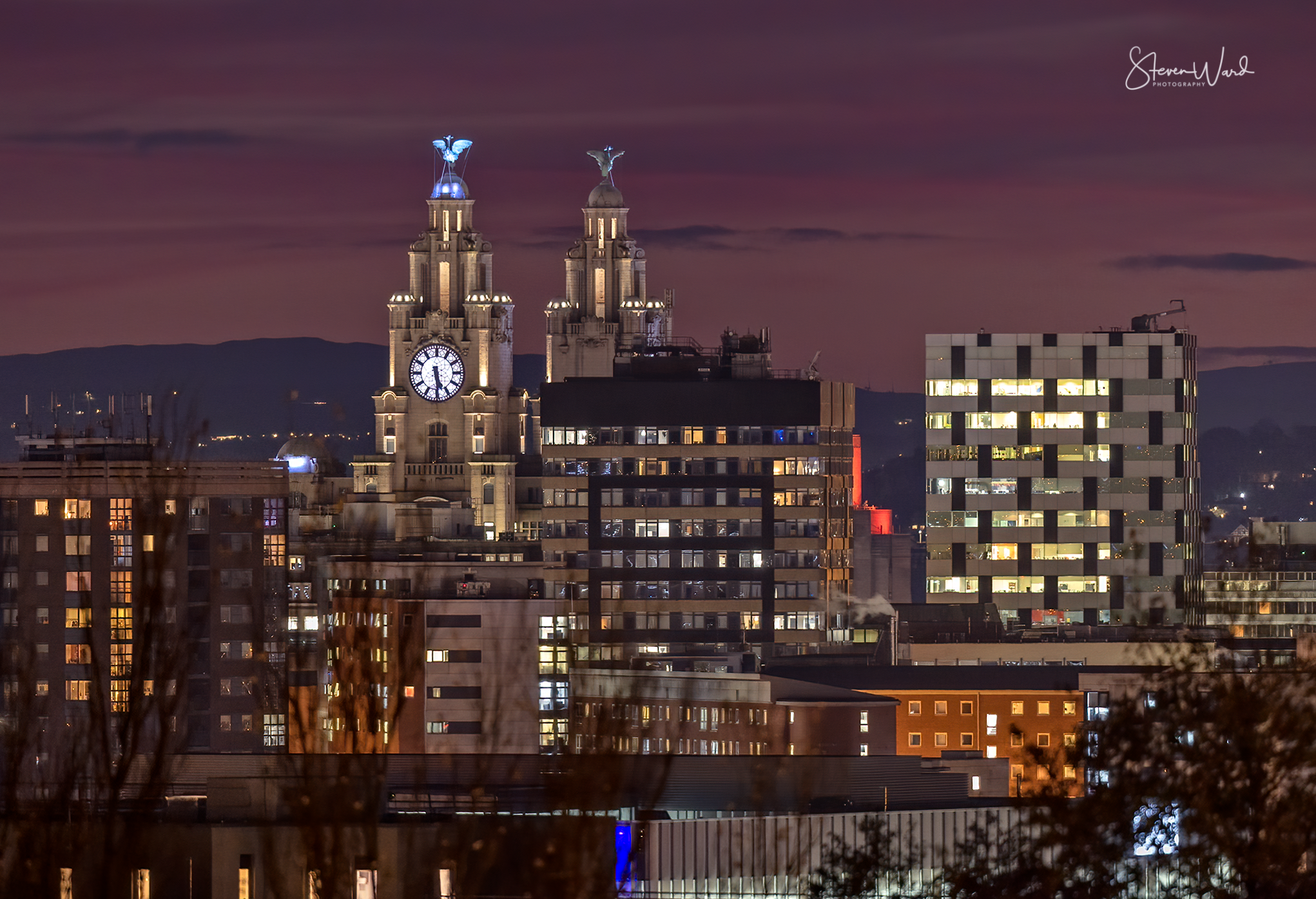 Nighttime city skyline featuring illuminated buildings and a prominent clock tower with two statues on top, under a purple sky.