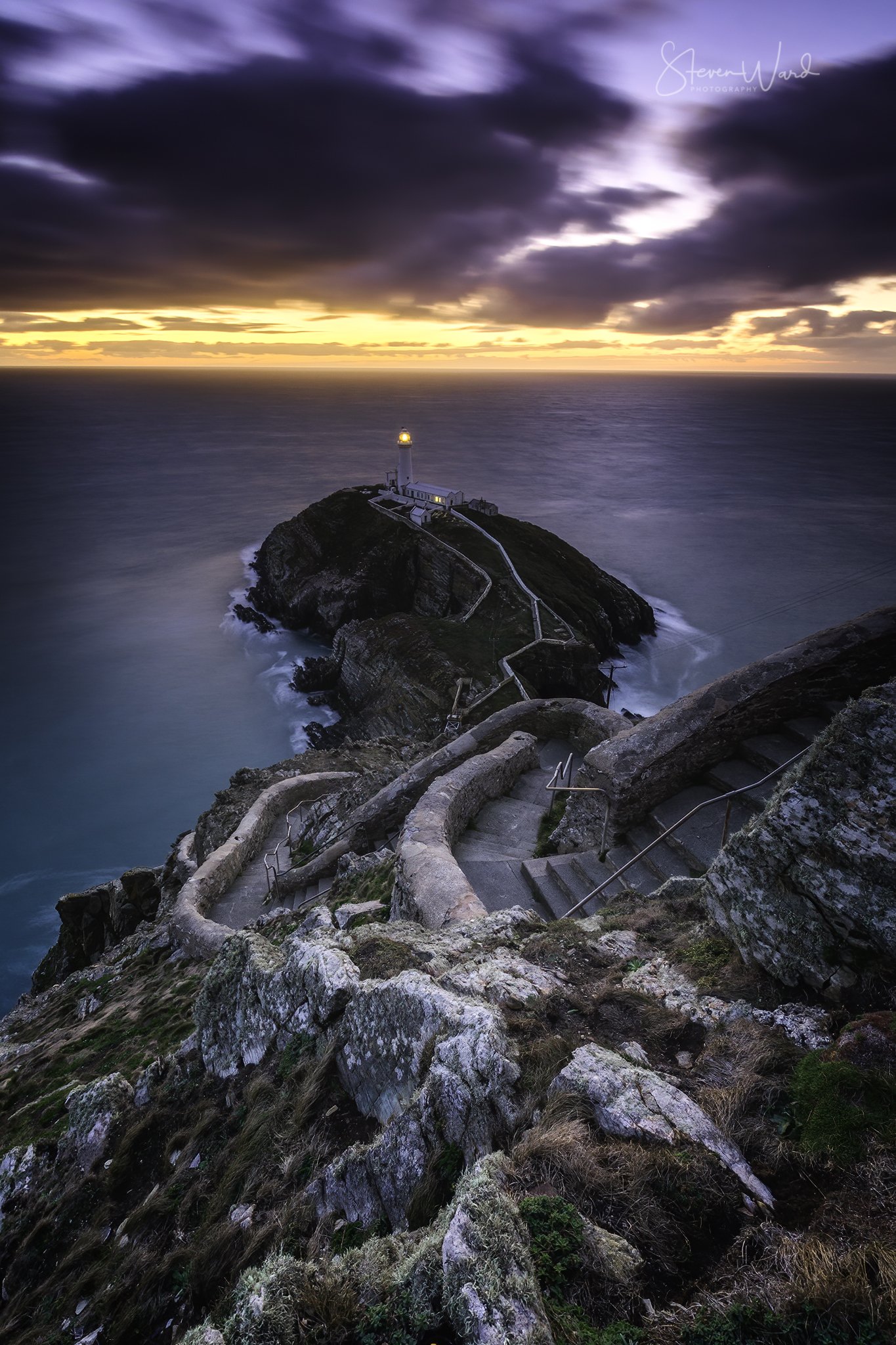 A winding staircase leading down a cliffside to a lighthouse on a rocky island at sunset, with dark clouds overhead and the ocean surrounding the island.