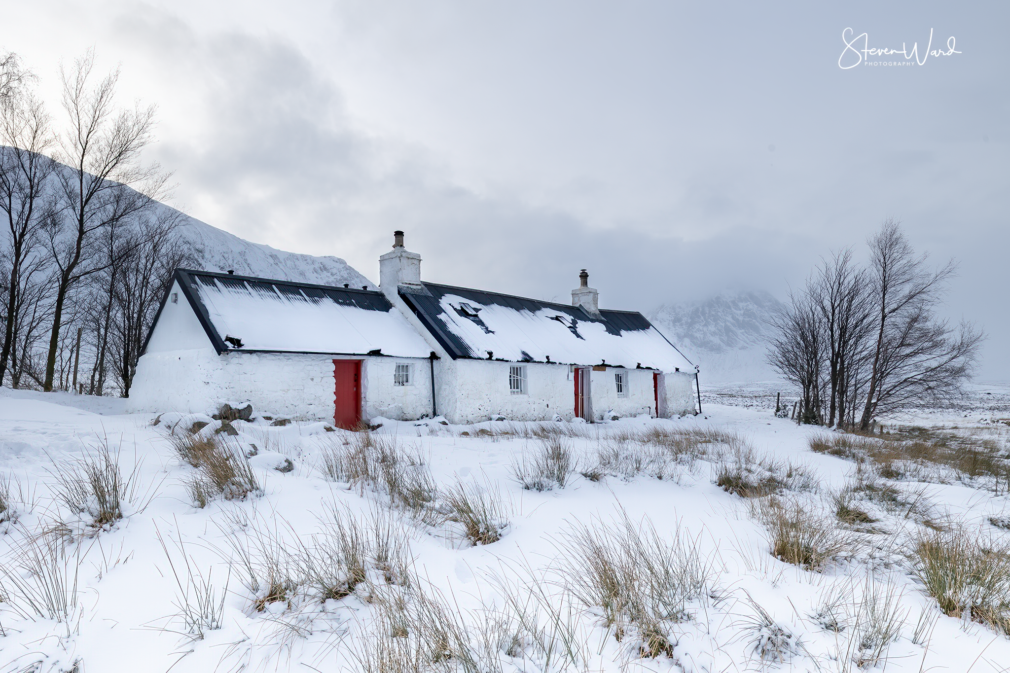 A white house with a black metal roof covered in snow, red doors, and two chimneys, set in a snowy landscape with bare trees and mountains in the background.