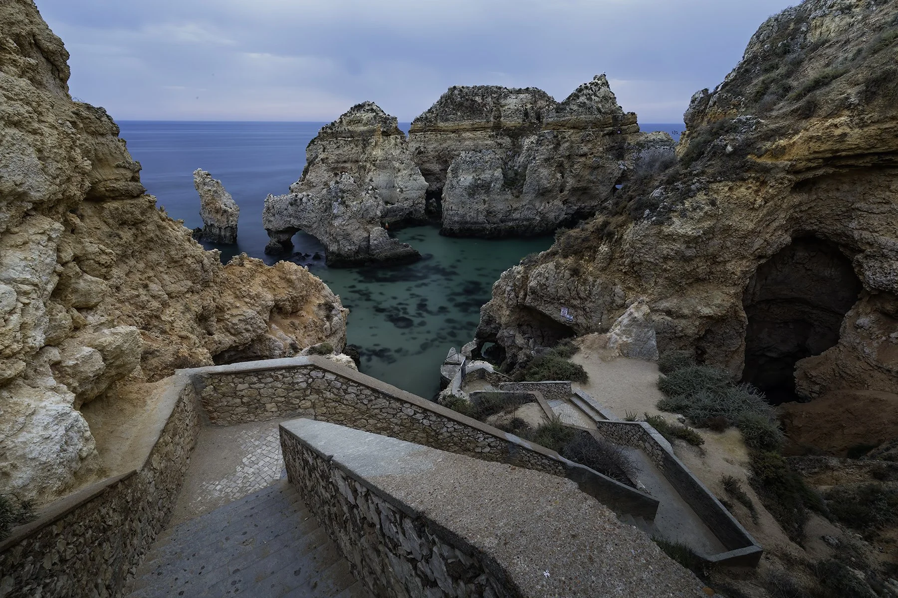 Photo of rocky coastline with pathways leading down to a small cove with turquoise water, surrounded by cliffs and rock formations under cloudy sky.