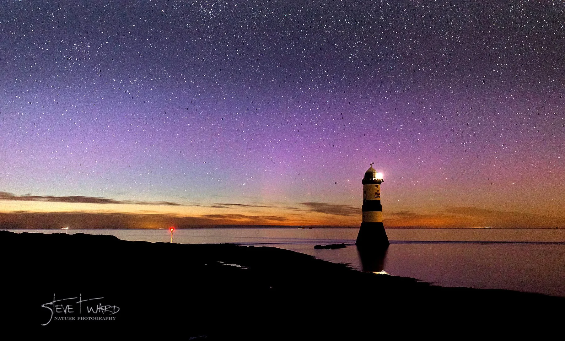 A lighthouse by the water at night with a starry sky overhead and a distant sunset on the horizon.