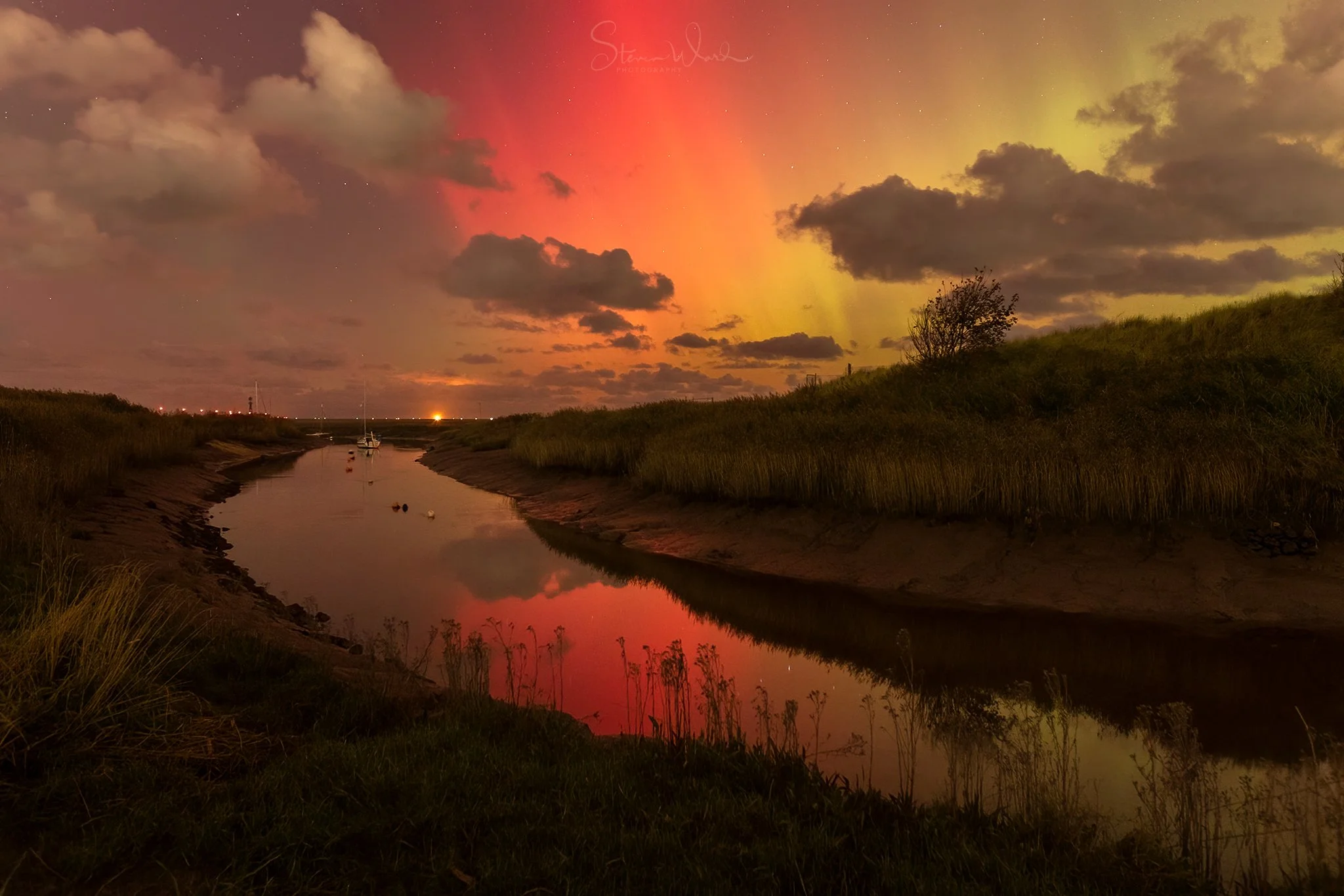 A small river or canal with still water reflecting a colorful sky at sunset or sunrise, with a mix of clouds and the aurora borealis in shades of pink, orange, and yellow, bordered by grassy banks and a small bush on the right.
