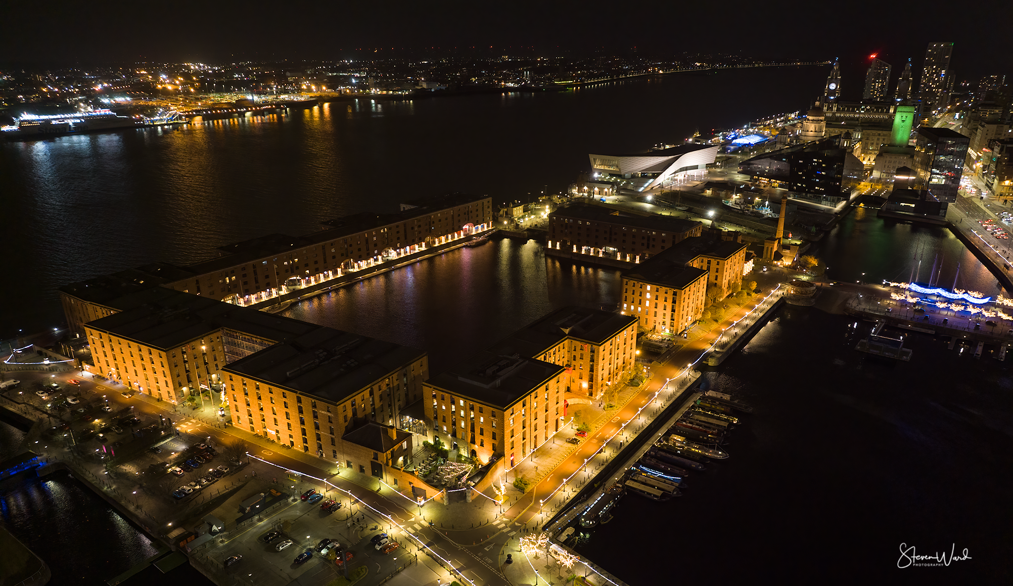 Aerial night view of a city with buildings, water, and illuminated streets, including a prominent U-shaped building complex and a river with boat reflections.