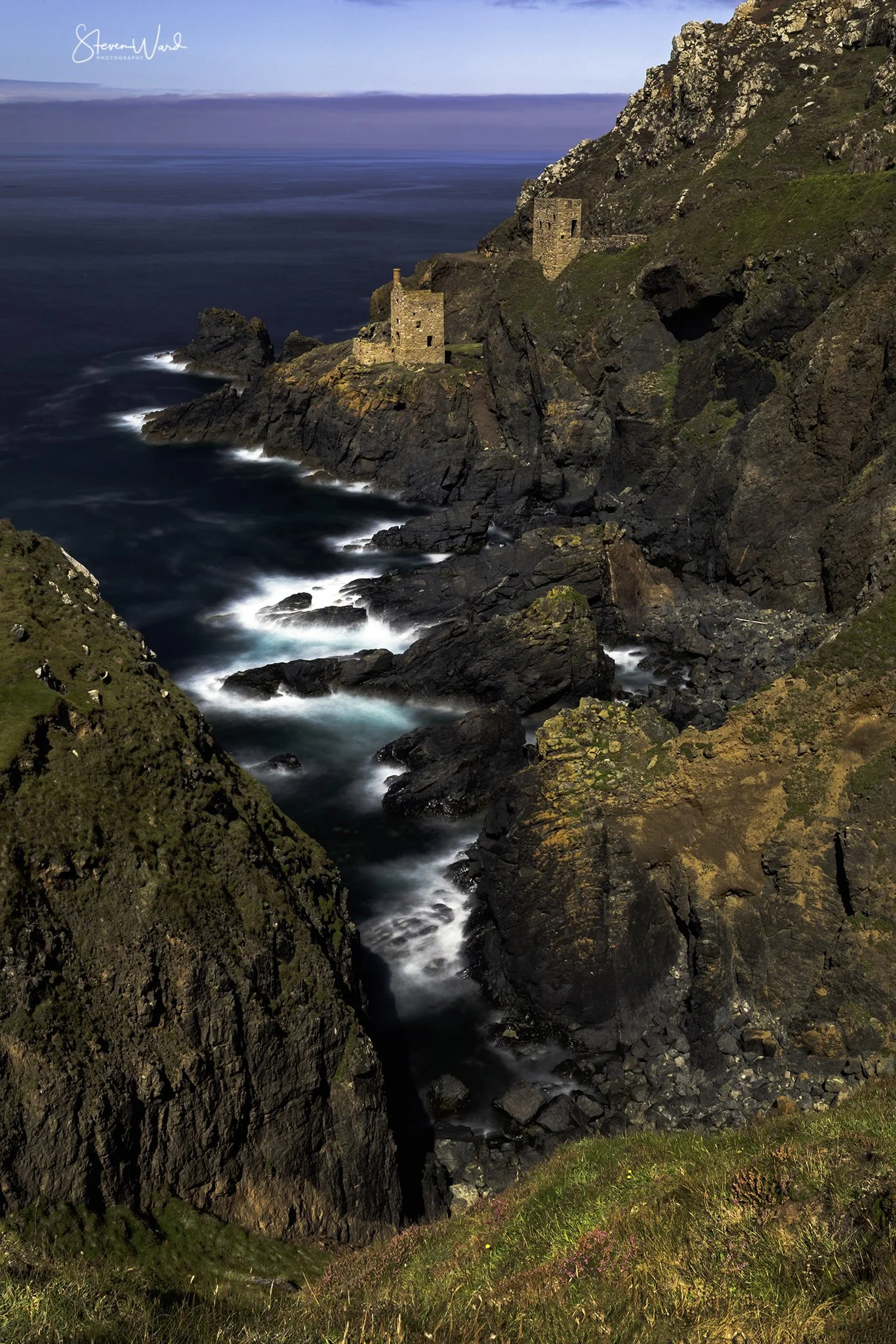Coastal cliffs with rocky terrain, green vegetation, and two stone ruins overlooking the ocean, under a partly cloudy sky.