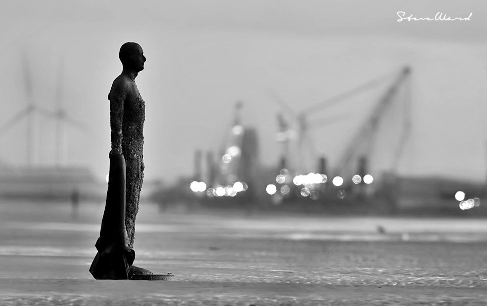 A silhouette of a person standing alone on a flat surface, with blurred industrial cranes and wind turbines in the background, in black and white.