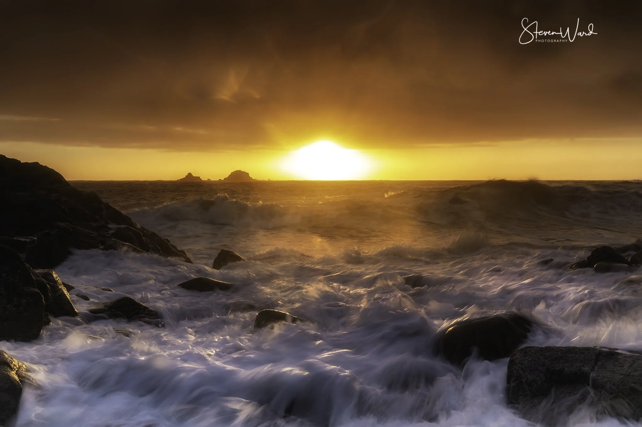 Sunset over the ocean with waves crashing against rocks in the foreground, dark clouds overhead.