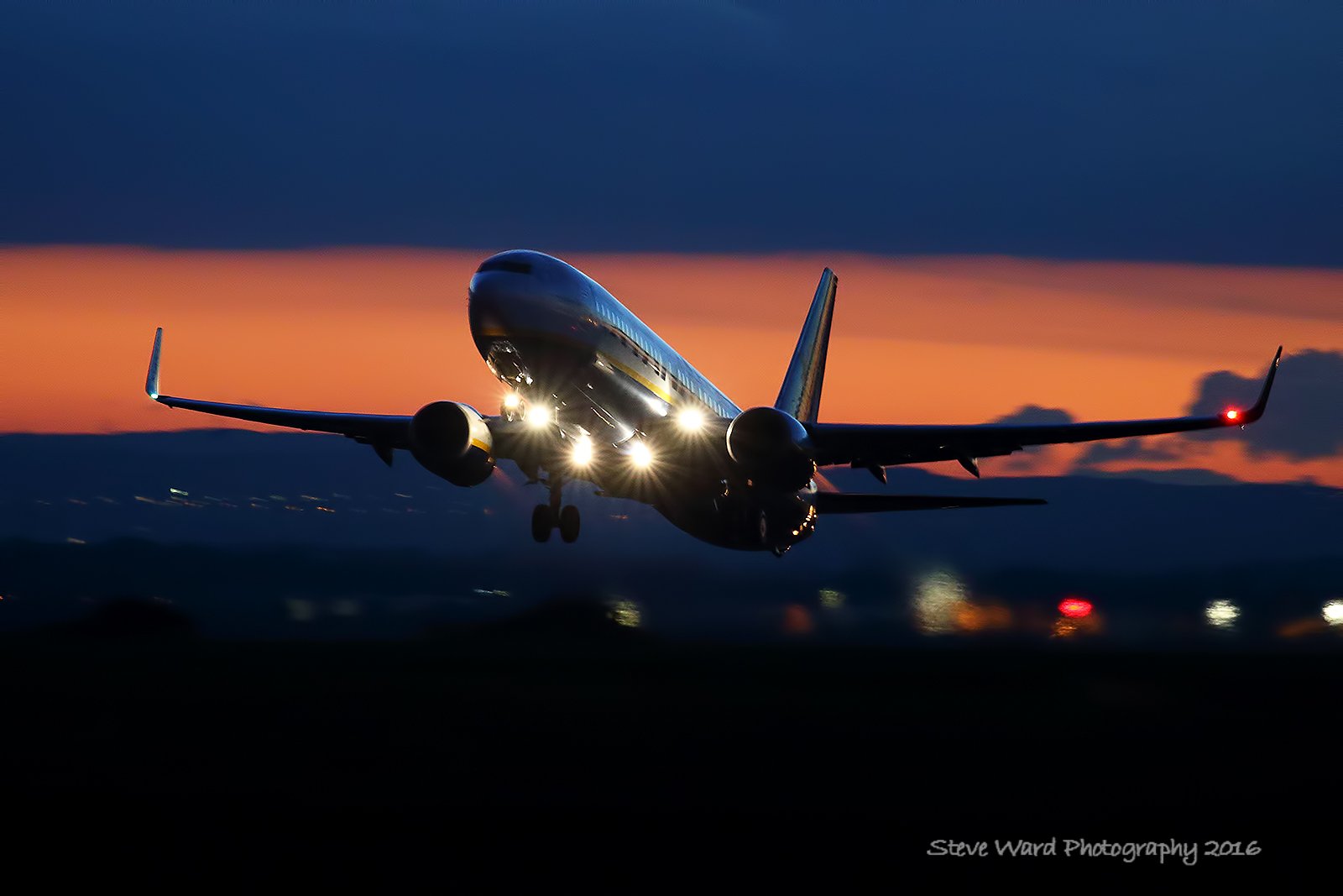 An airplane taking off during sunset with landing lights on, dark sky with orange and blue hues, and the silhouette of the landscape below.