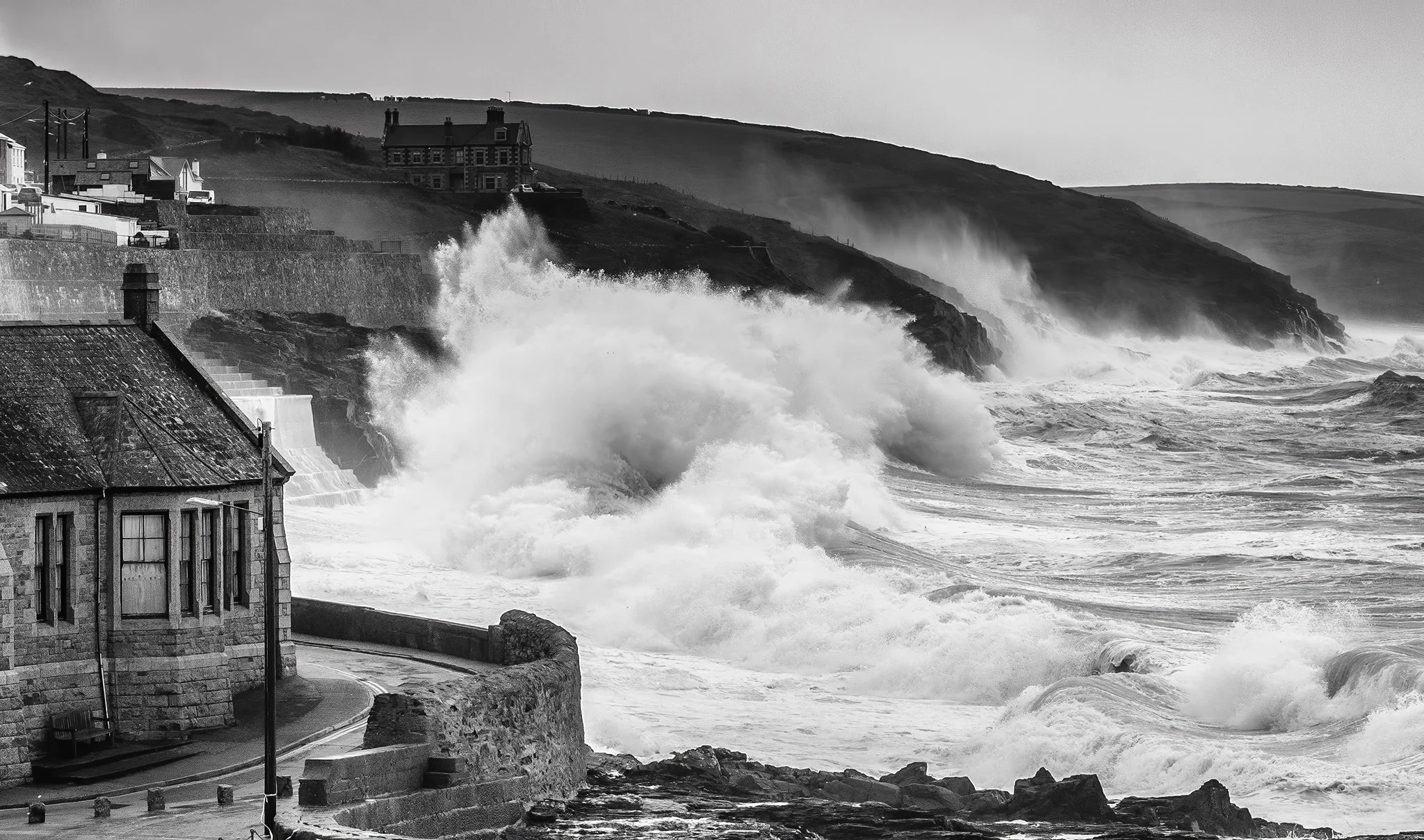 Black and white photo of large ocean waves crashing against a rocky shoreline with houses on a hill in the background.