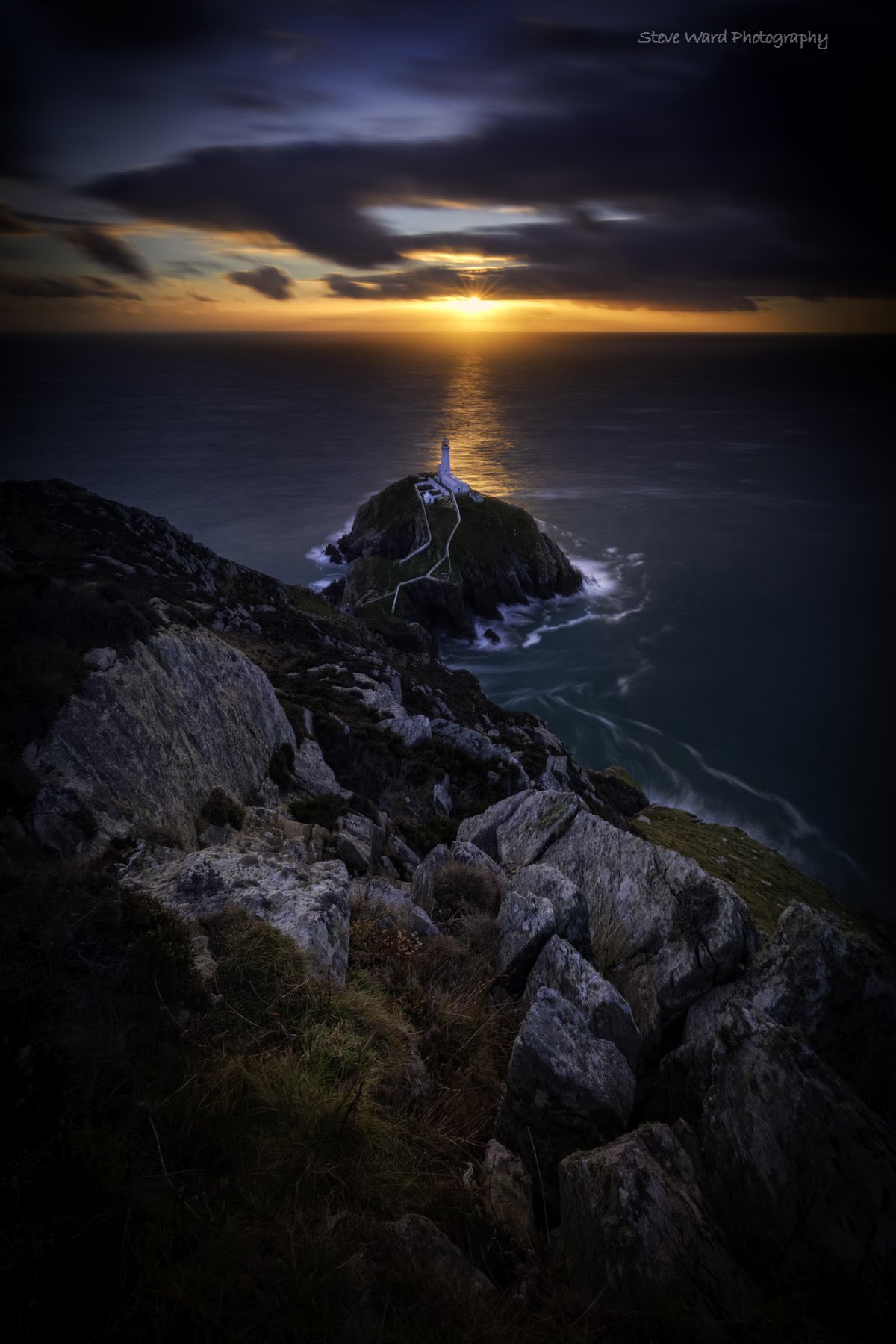 Sunset over the ocean with a lighthouse on a rocky island connected by a pathway, viewed from a hillside with rocks and grass in the foreground.