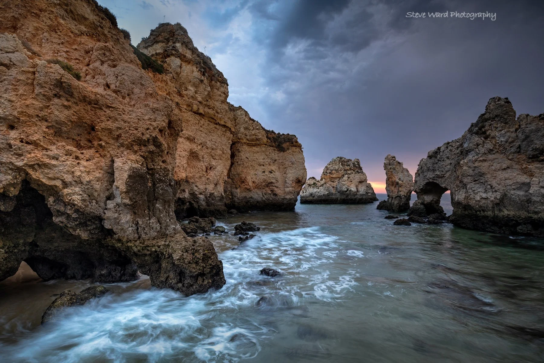 Rocky cliffs by the ocean at sunset with waves crashing on the shore and a cloudy sky.