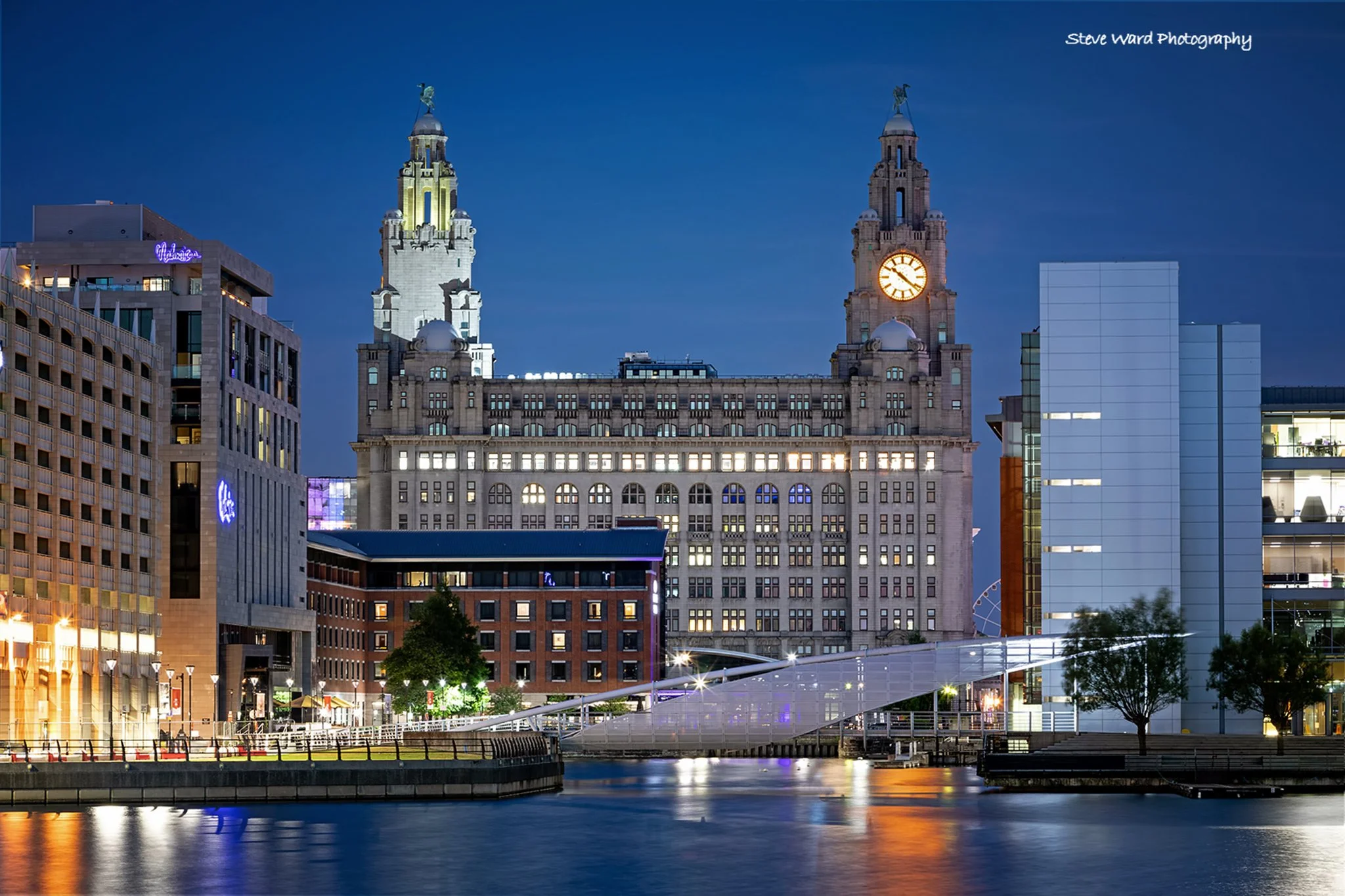 Nighttime cityscape featuring historic and modern buildings, illuminated clock tower, and a waterway in Liverpool, England.