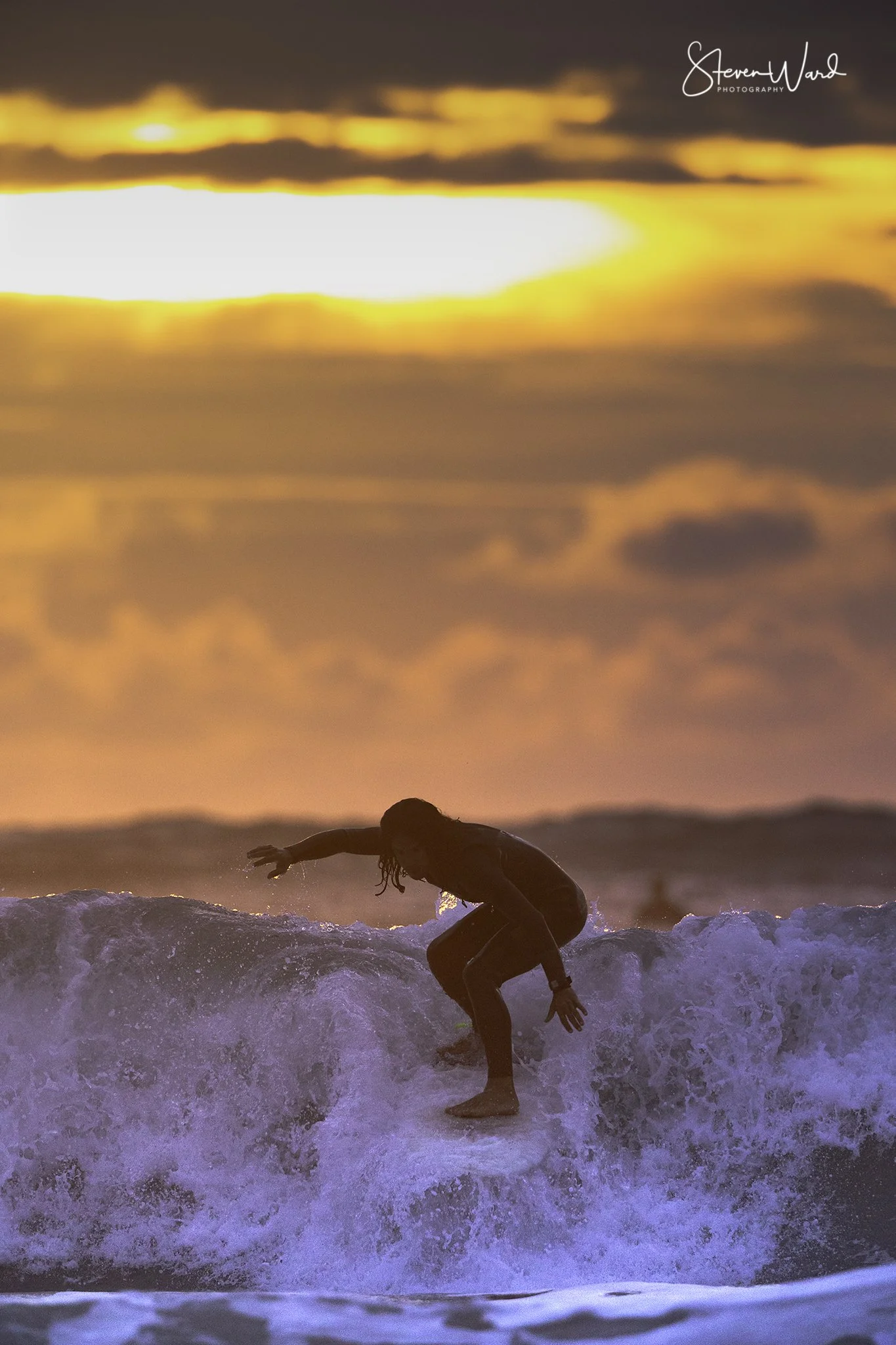 A person with long hair surfing on a wave during a vibrant sunset, with orange and pink clouds in the sky.