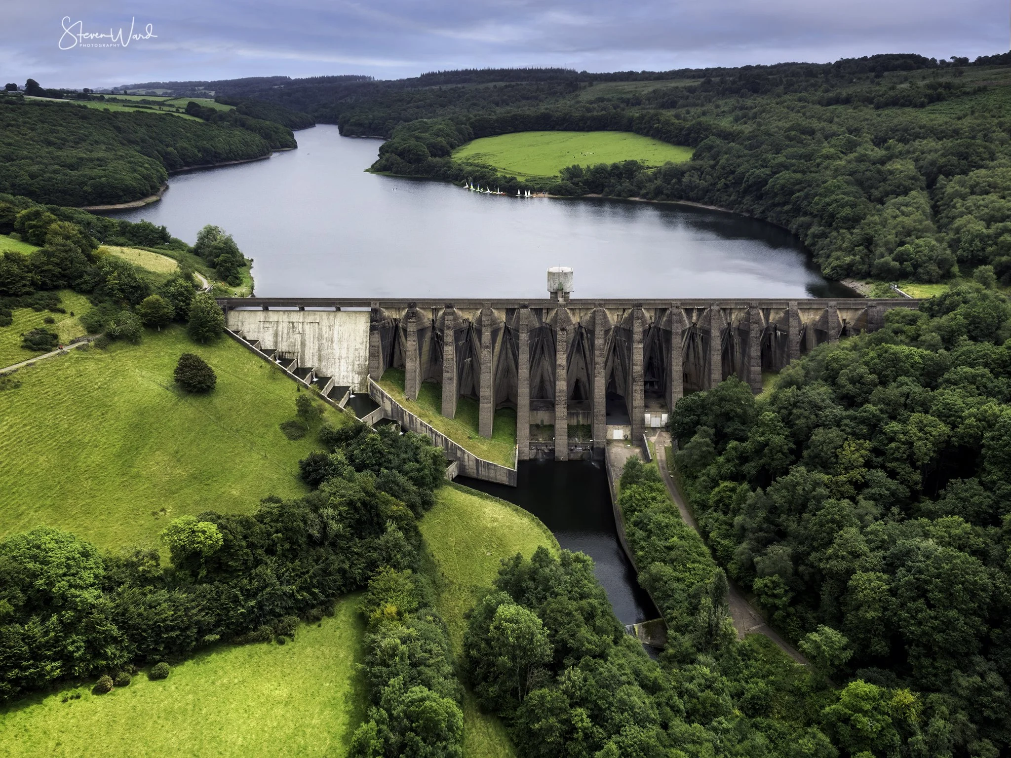 An aerial view of a large dam surrounded by lush green trees and rolling hills, with a reservoir behind it and water flowing through the dam's spillway.