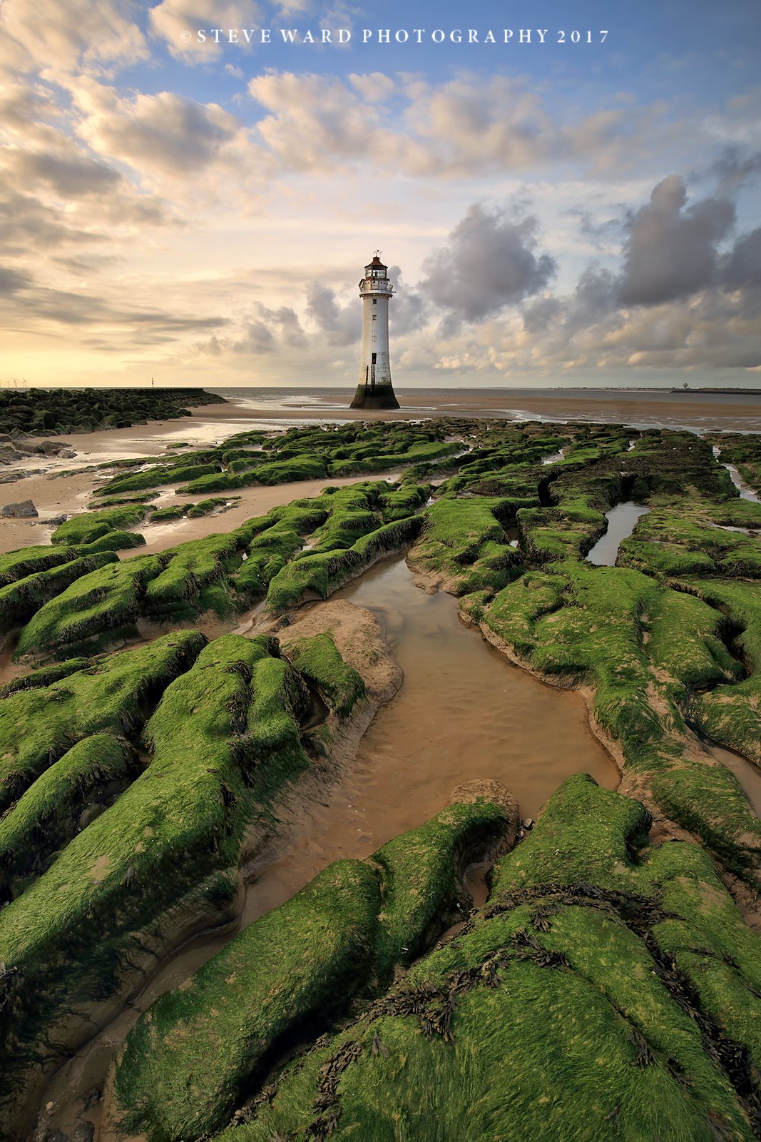 A lighthouse standing on a sandy beach with green moss-covered rocks and tide pools in the foreground, under a partly cloudy sky at sunset.