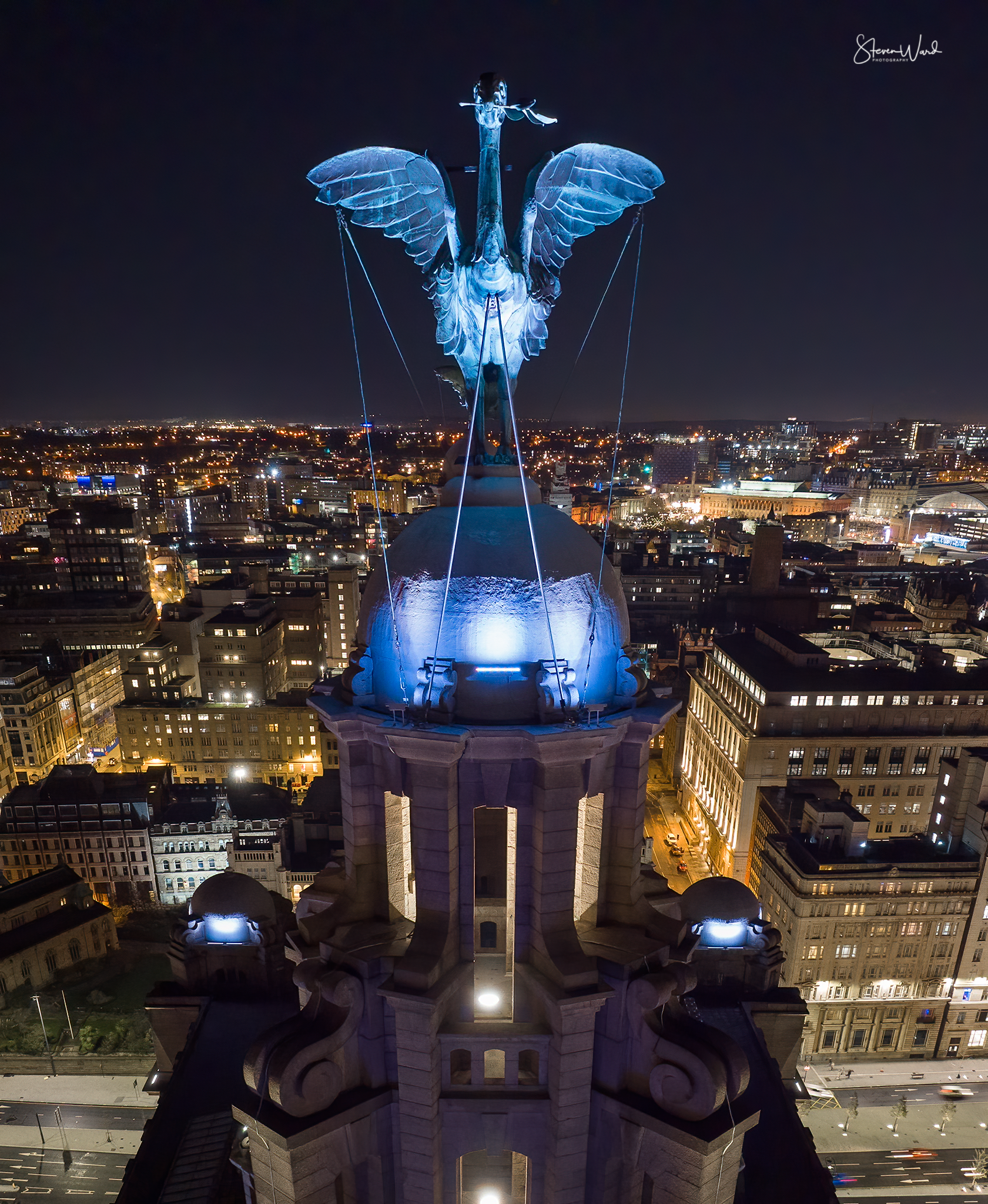 Nighttime aerial view of the top of a historic building with an illuminated angel statue on the roof, overlooking a cityscape with lights and buildings in the background.