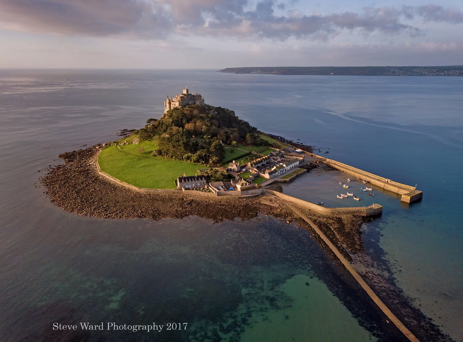 Aerial view of a small island with a castle on top, surrounded by water, with a harbor and small boats on the right side, and a green lawn and stone walls on the island.