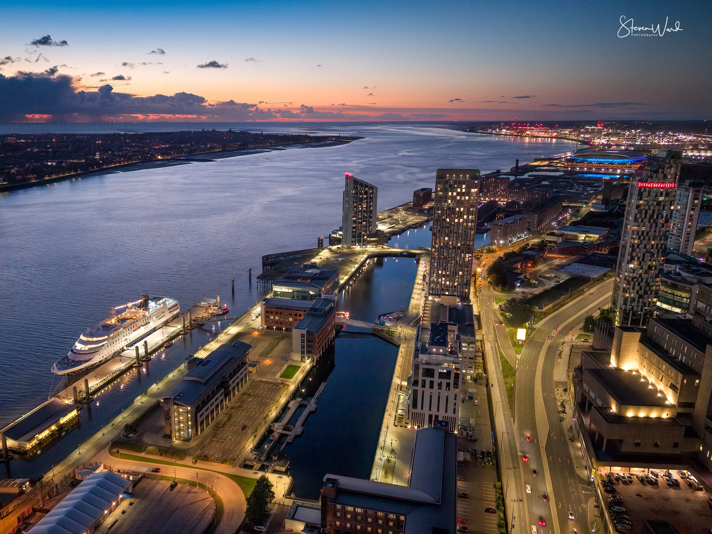 Aerial view of a city harbor at dusk with illuminated buildings, boats, and a river flowing through the city.