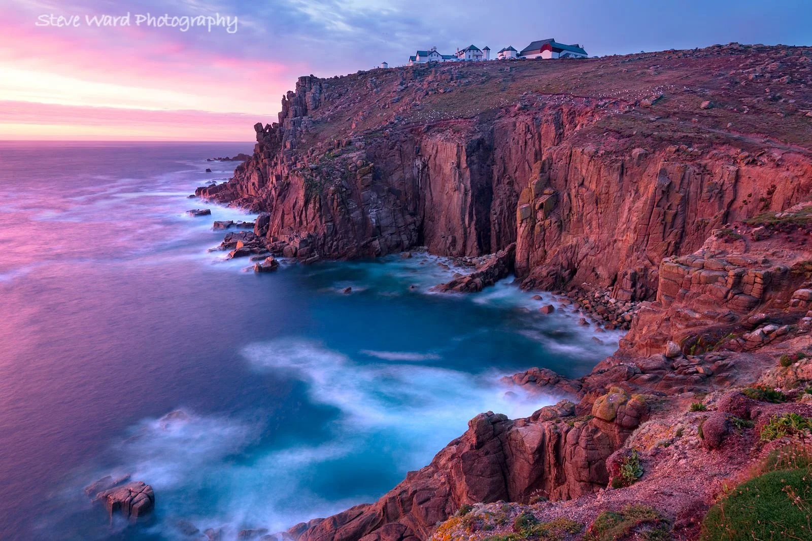 Colorful sunset over rocky coastal cliffs with white houses on top, and ocean waves crashing below.