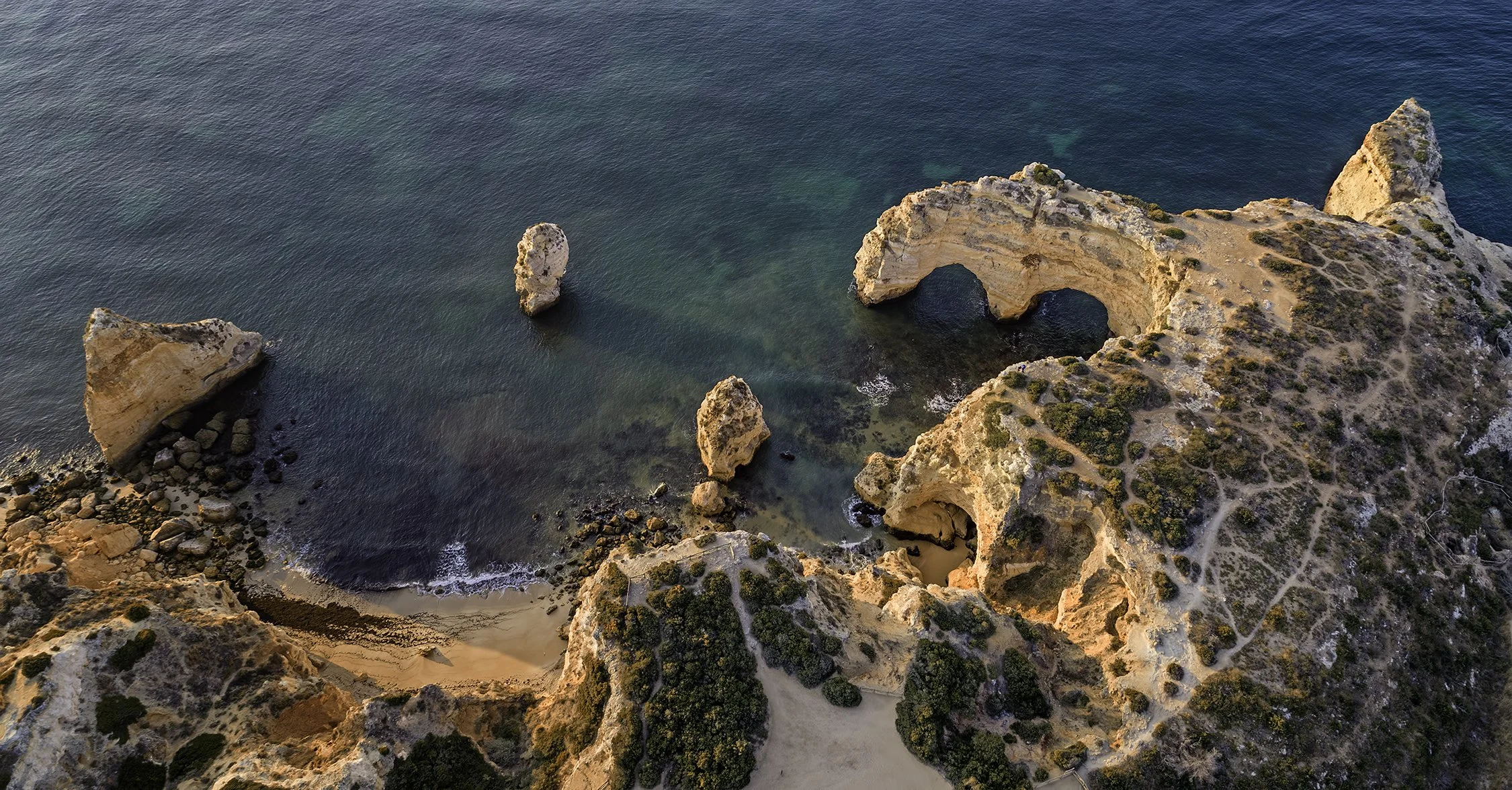 Aerial view of a coastal landscape with rugged cliffs, sea stacks, and a natural arch formation over the ocean, with sandy beach and pathways.