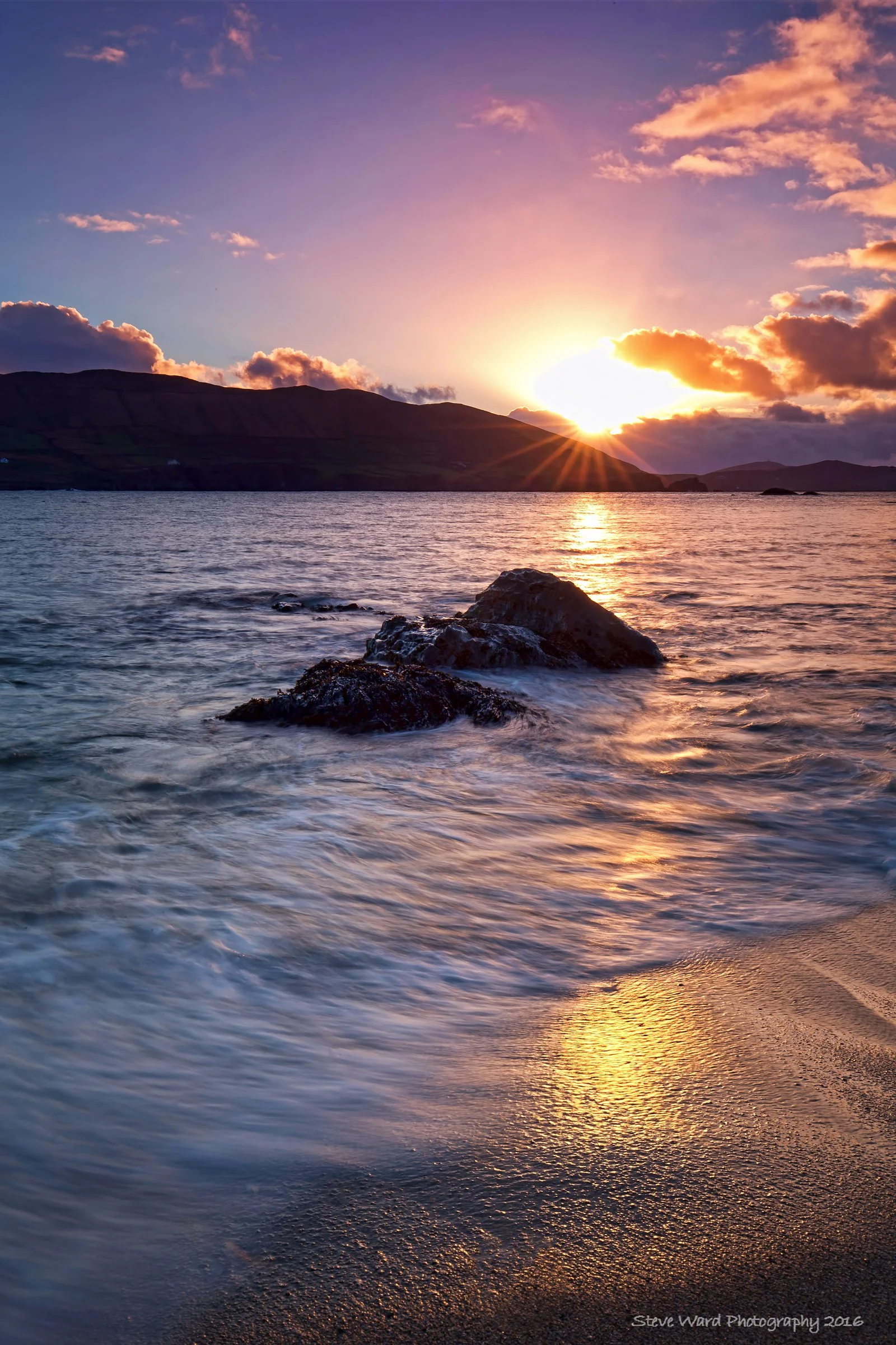 A beautiful sunset over the ocean with colorful clouds, visible rocky shoreline, and reflected sunlight on the water and sand.